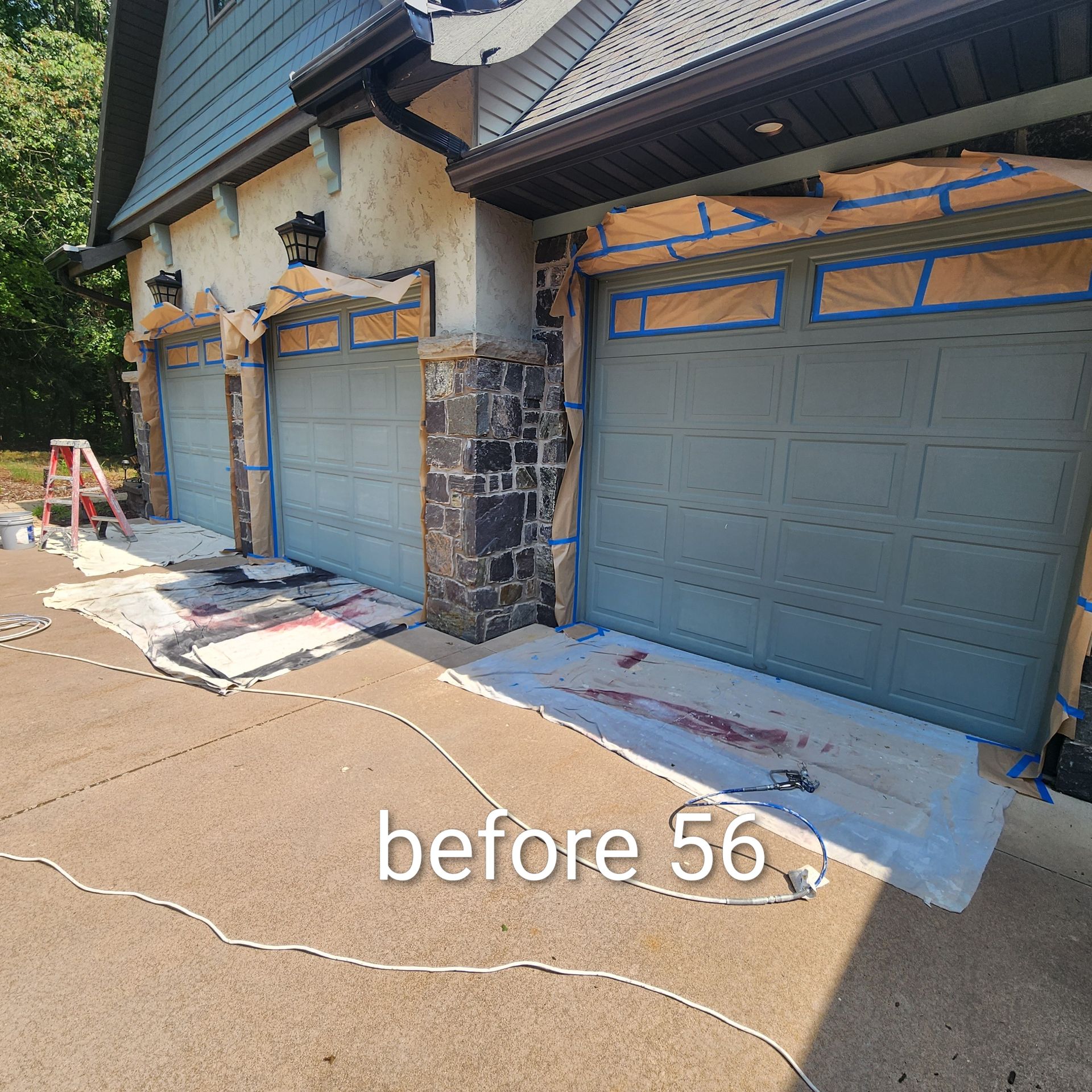 Two garage doors on a house exterior being painted gray with blue painter’s tape and drop cloths