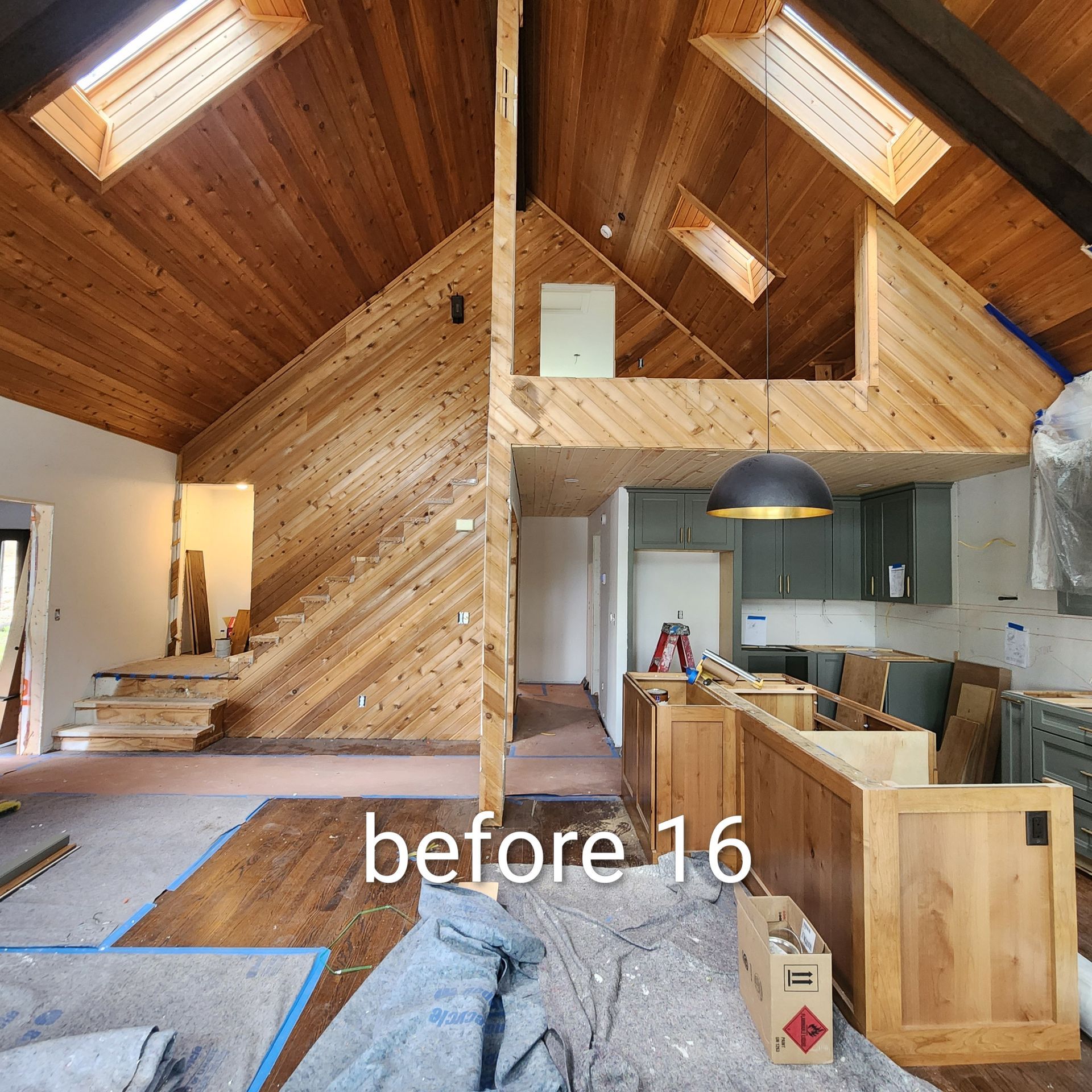 Wood-paneled A-frame interior under renovation, with exposed framing, kitchen cabinets, and stairway.