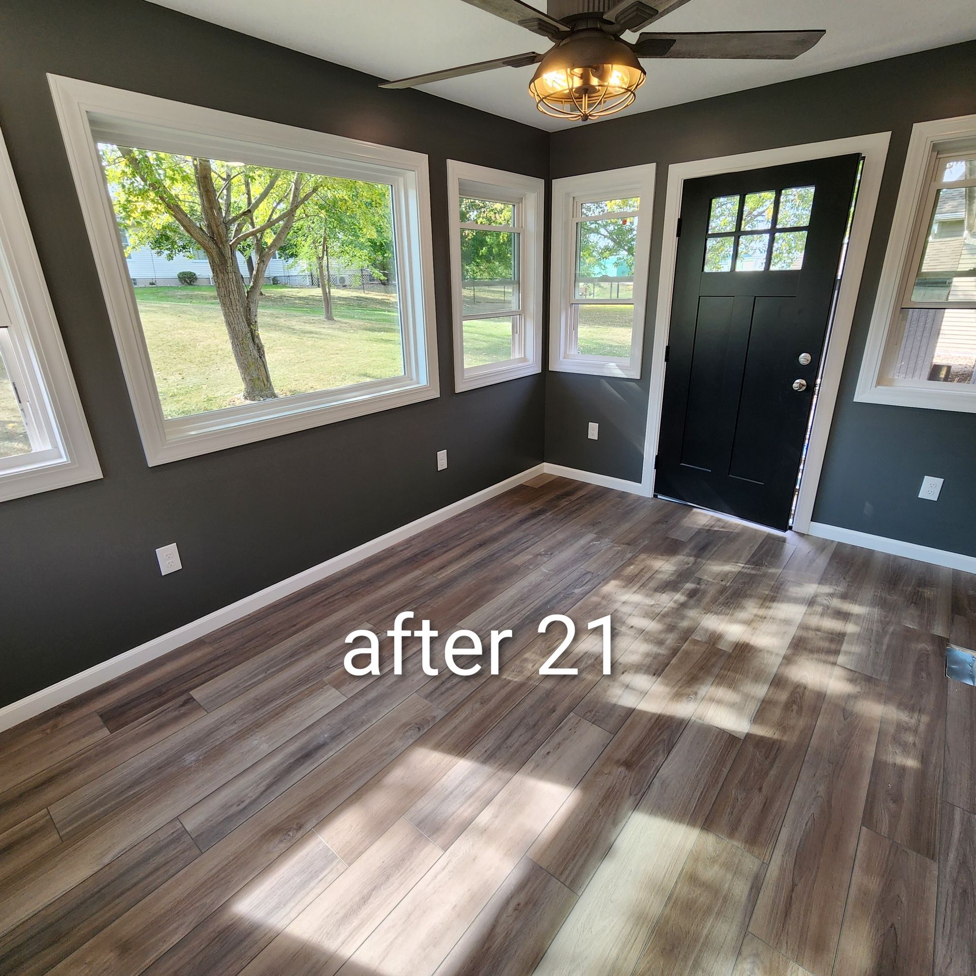 Empty room with dark walls, wood-look floor, large windows, and a black front door.