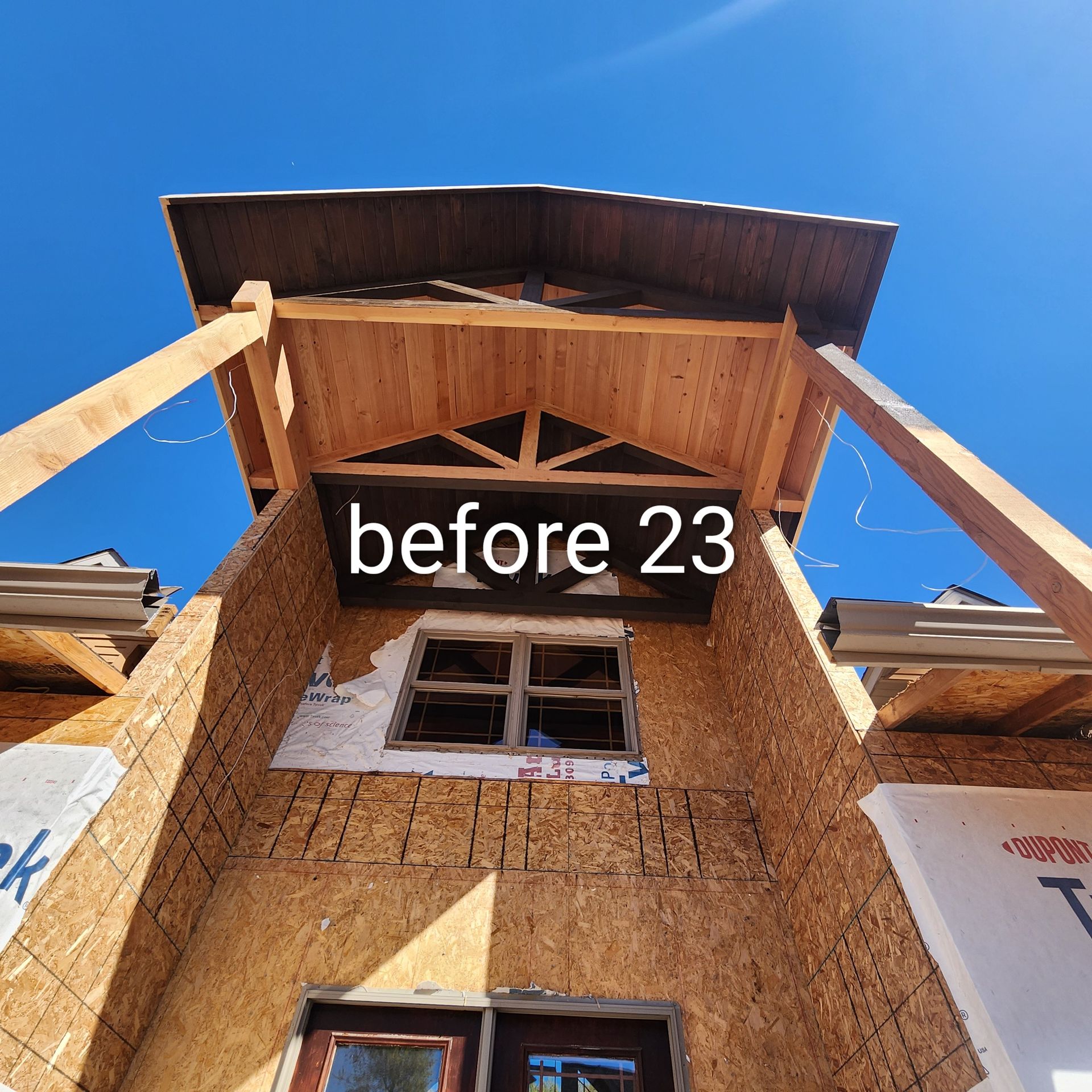 Wooden building frame under construction against a bright blue sky, with a window opening and “before 23” sign.