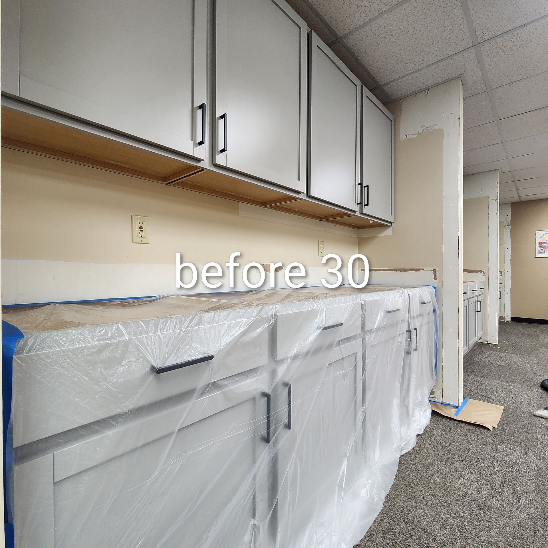 Office cabinets and countertop covered in plastic sheeting during renovation, with carpeted hallway and cleaning tools.