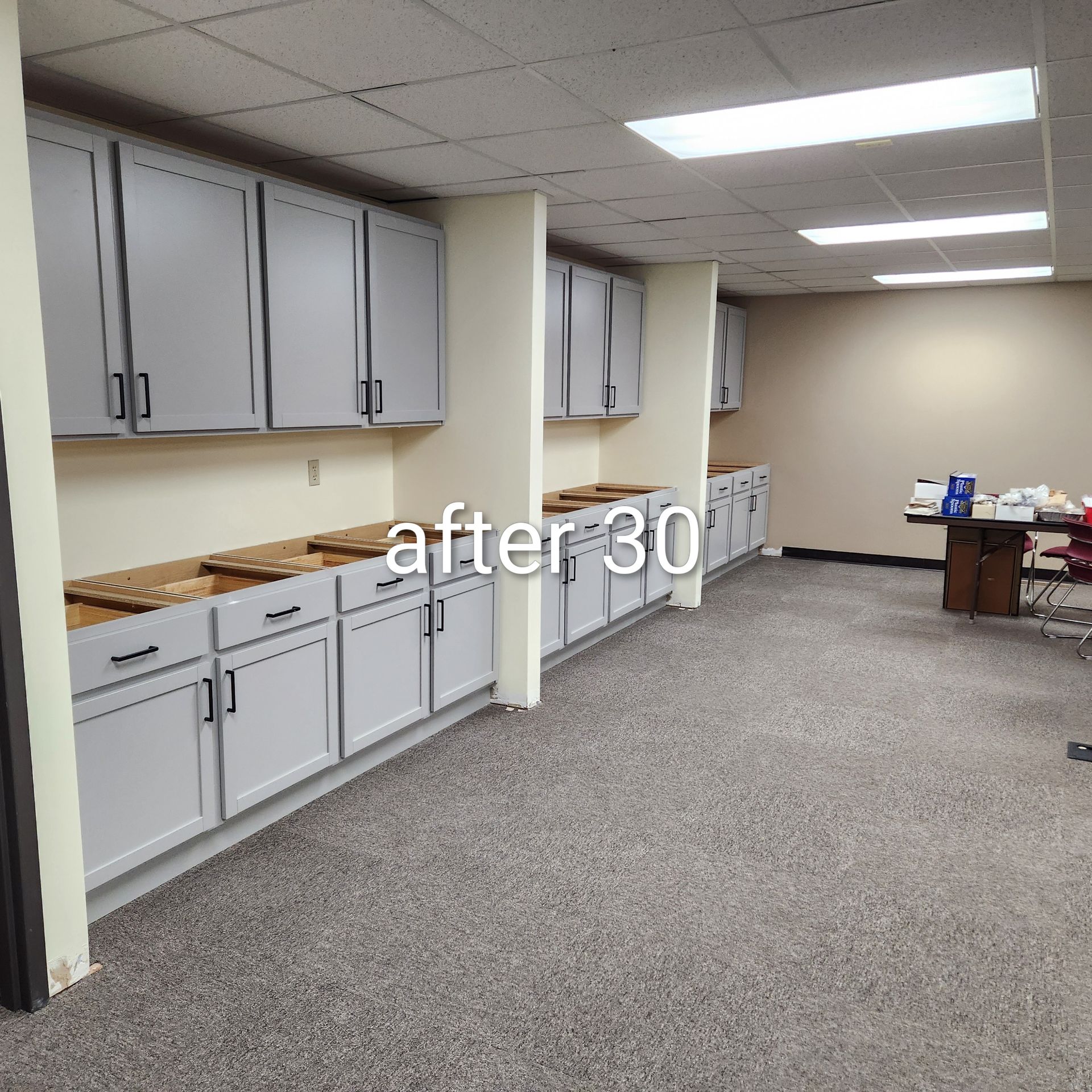 Empty office break room with gray cabinets, carpet, and fluorescent ceiling lights.