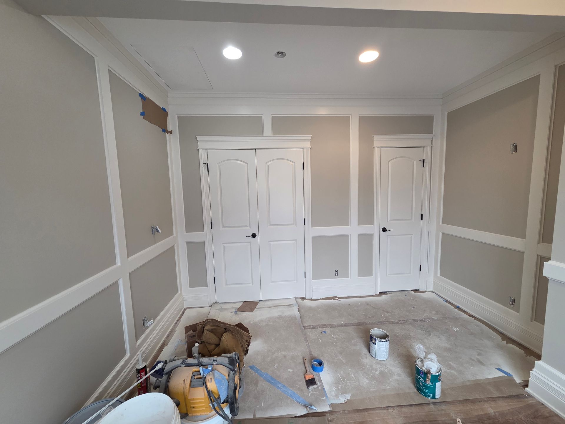 Empty room under renovation with white trim, beige walls, recessed lights, and tools and materials on the floor.