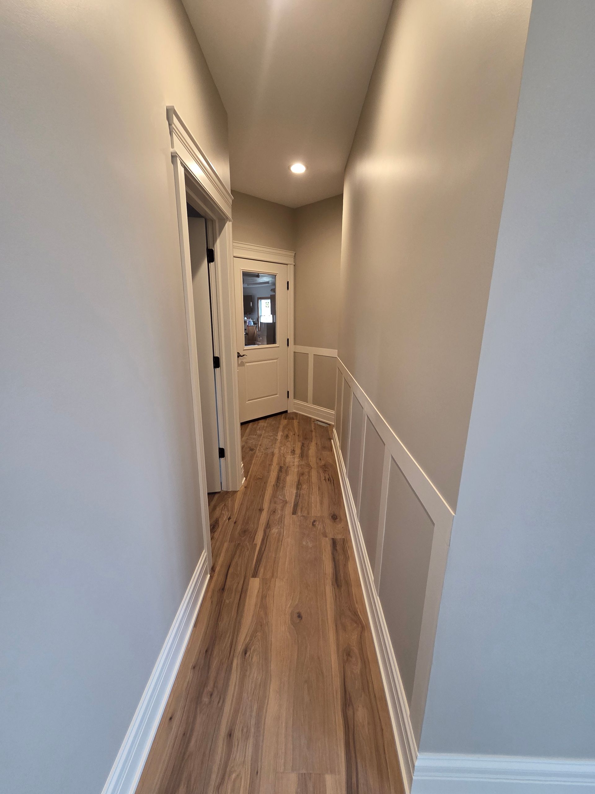 Narrow hallway with wood floors, beige walls, white wainscoting, and a small window at the far end.