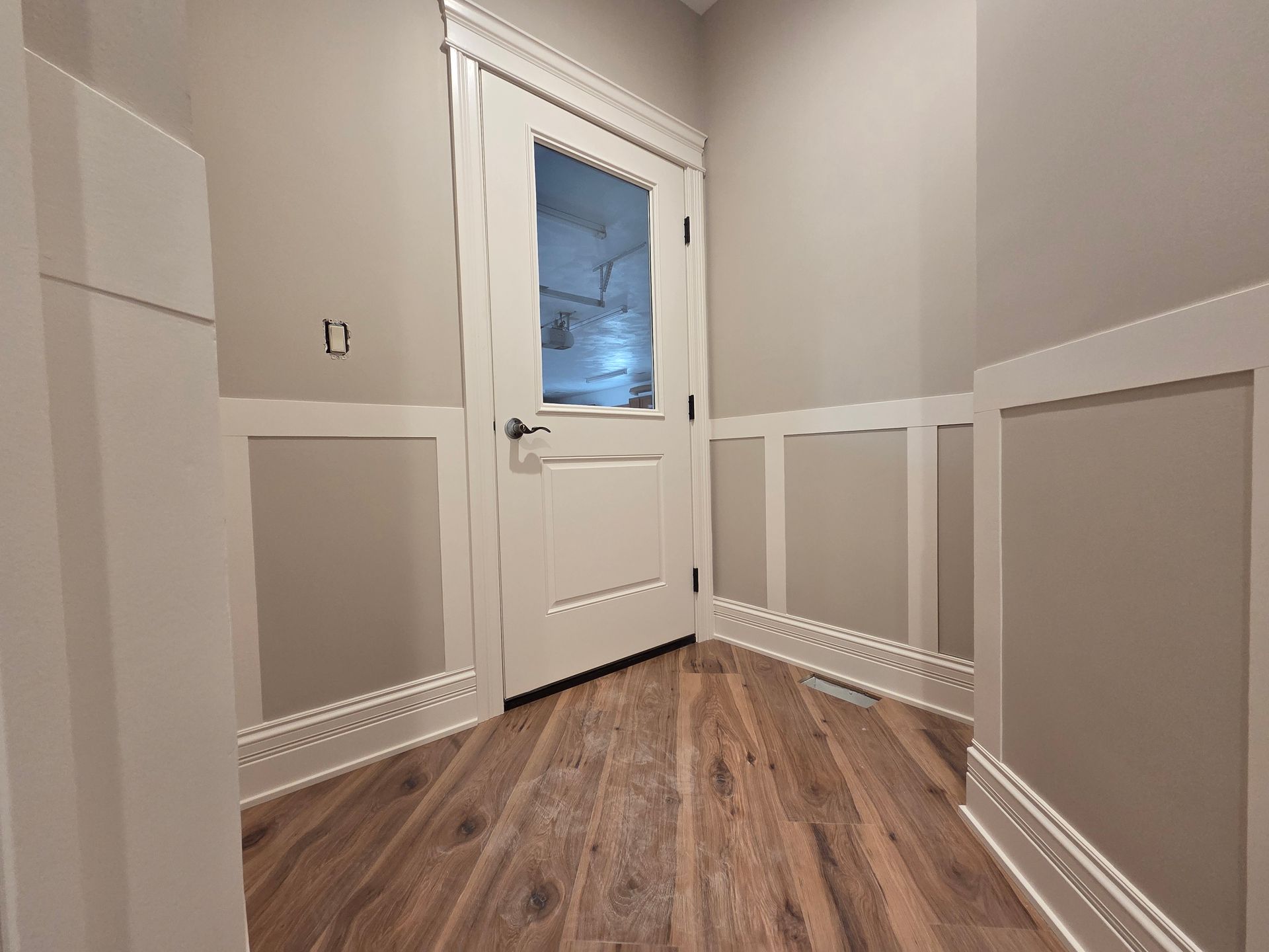 Narrow hallway with wood floor, white paneled walls, and a closed white door with a frosted window.