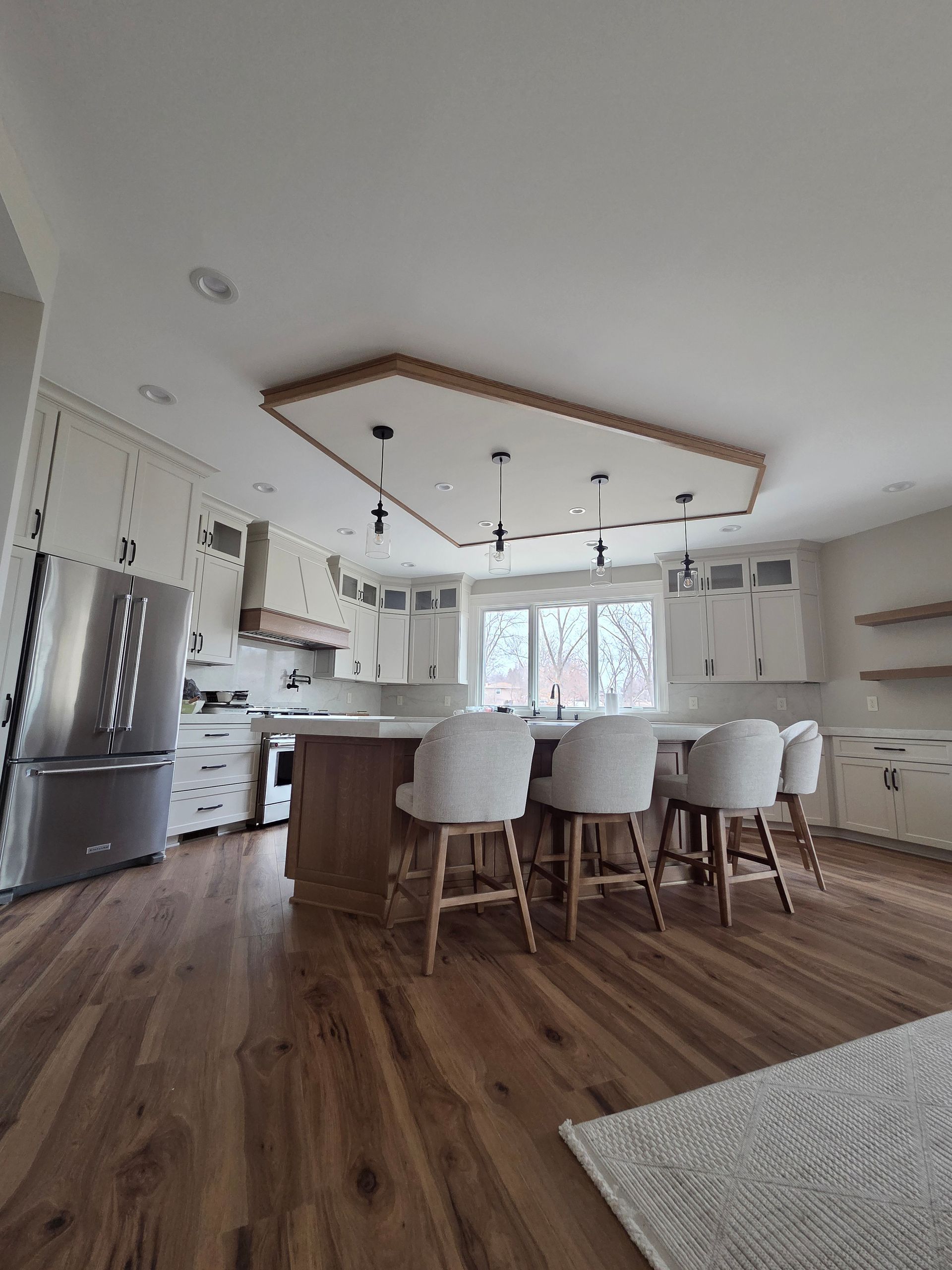Modern kitchen with island, white chairs, stainless fridge, wood floors, and a tray ceiling with pendant lights