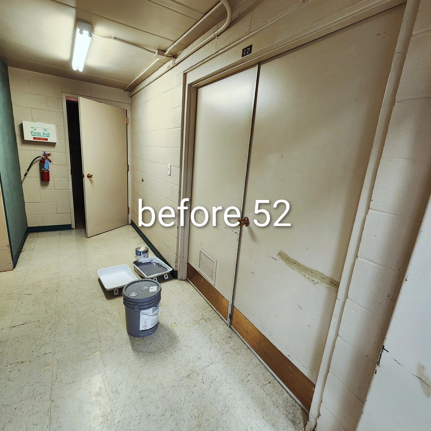 Hallway with white walls, closed double doors, and cleaning supplies on the floor under fluorescent lights