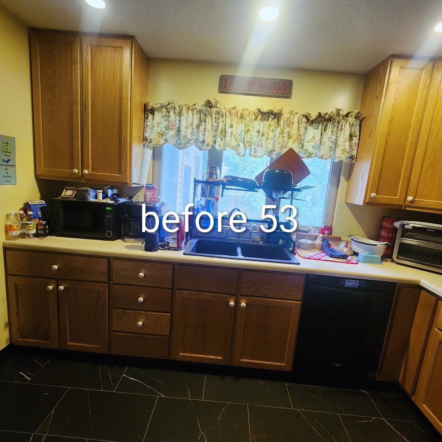 Kitchen with wooden cabinets, black dishwasher, and window over sink with floral curtains.