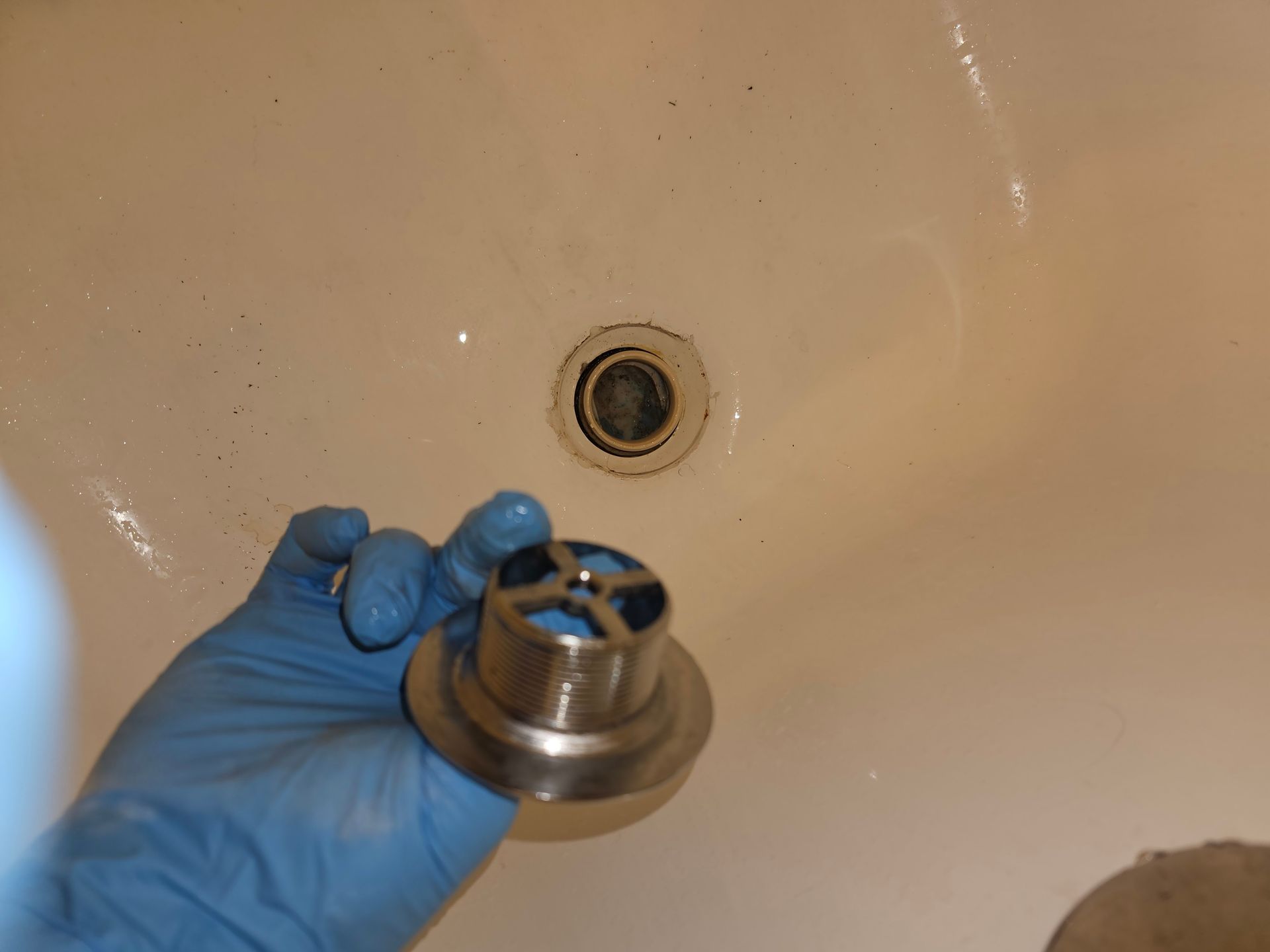 A gloved hand holds a new stainless steel bathtub drain cover next to the open drain in a white tub.