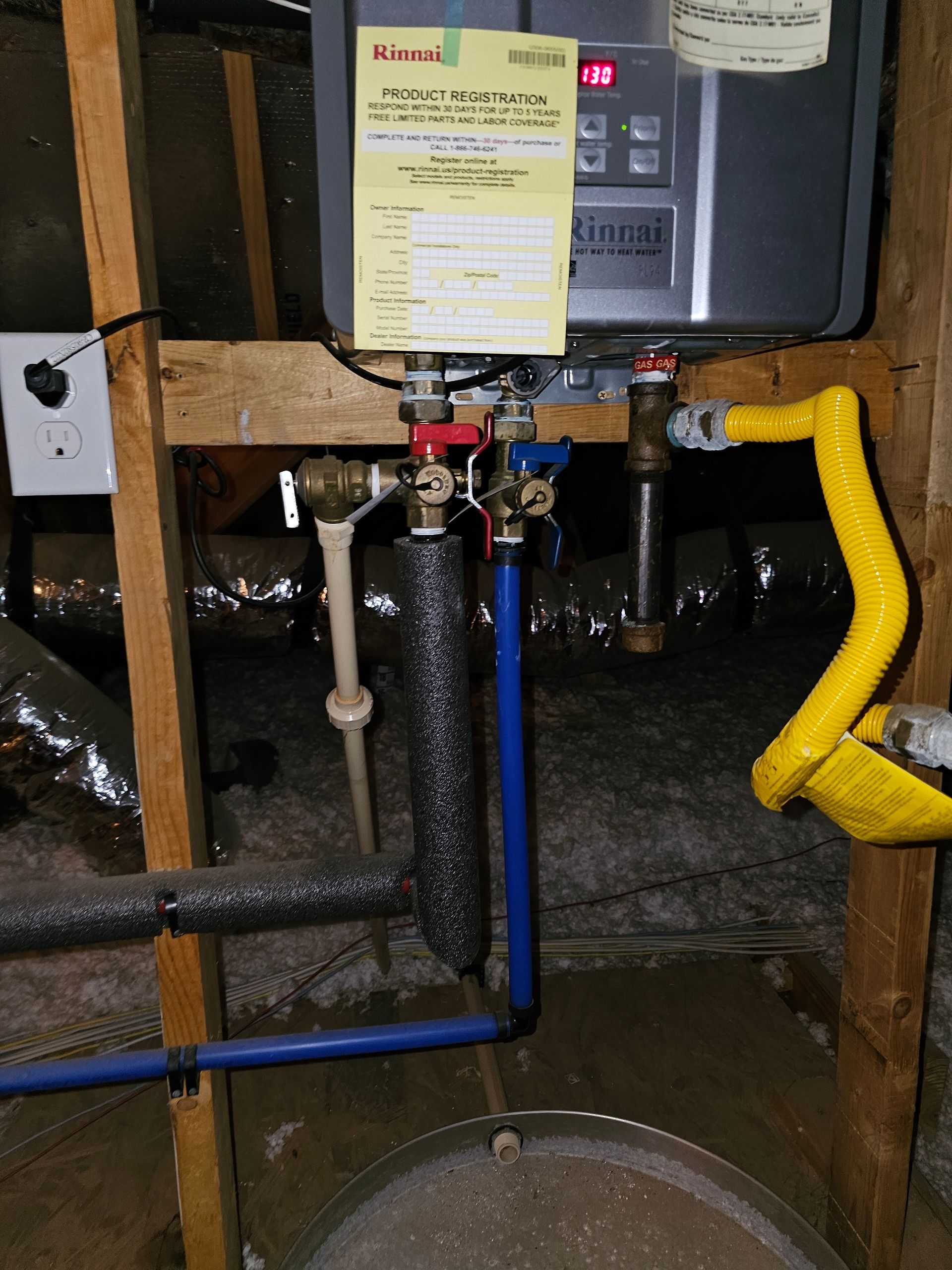 Tankless water heater with plumbing, electrical outlet, and pipes in an unfinished space.