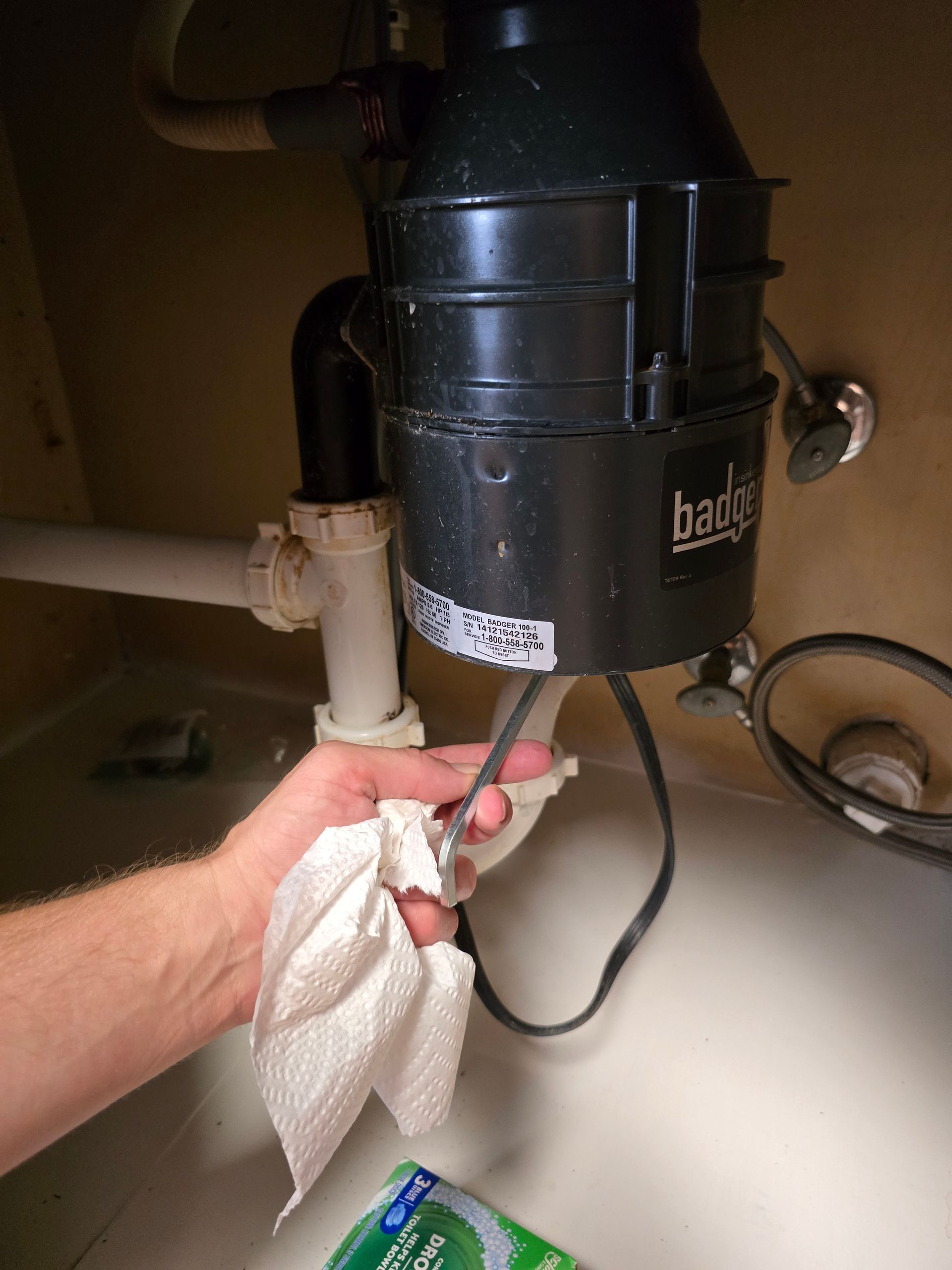 Person's hand holds a wet cloth near a garbage disposal unit under a sink. Black disposal unit has a power cord.
