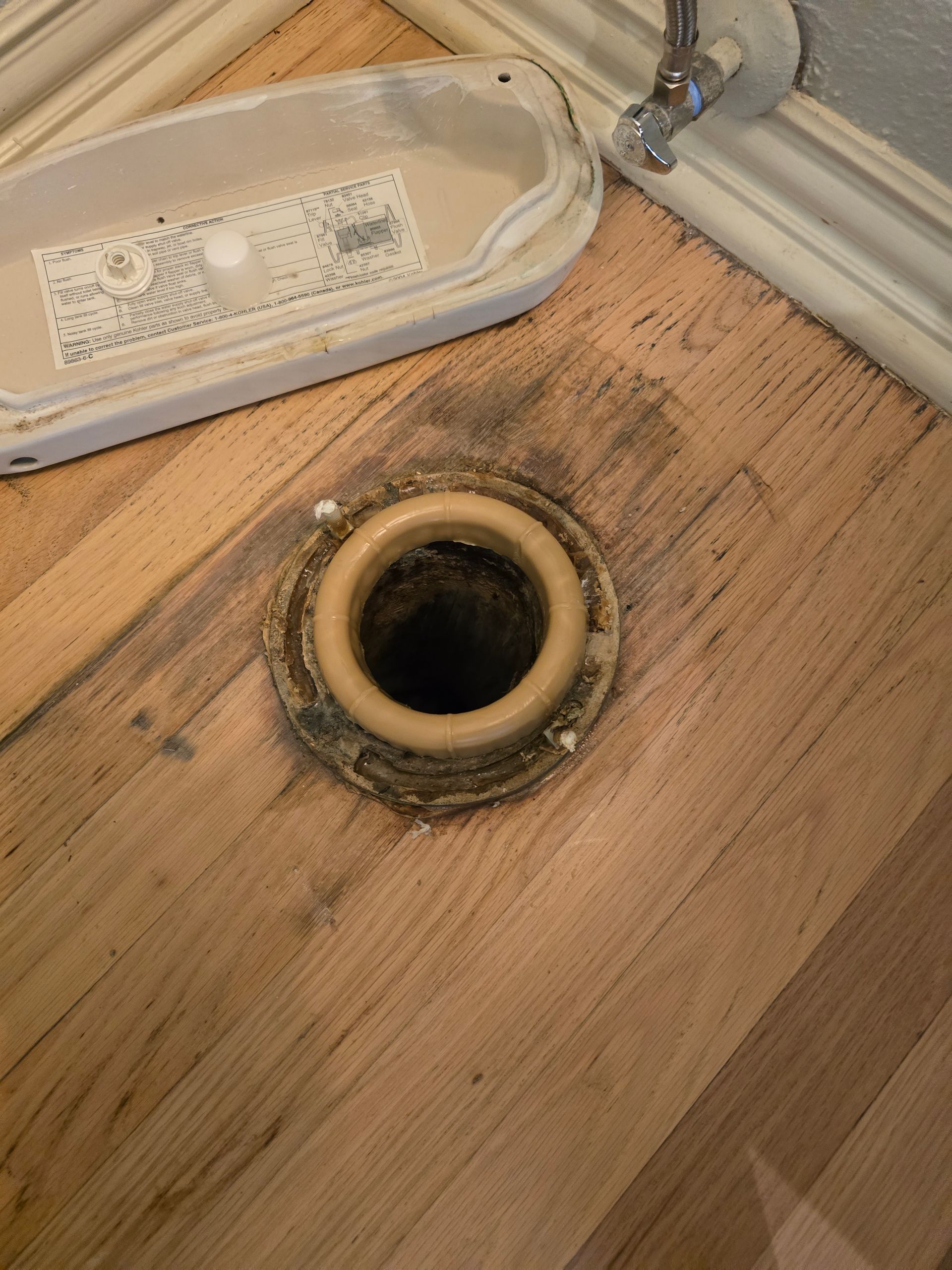 Toilet flange and drain opening on wooden floor, toilet tank lid is nearby.