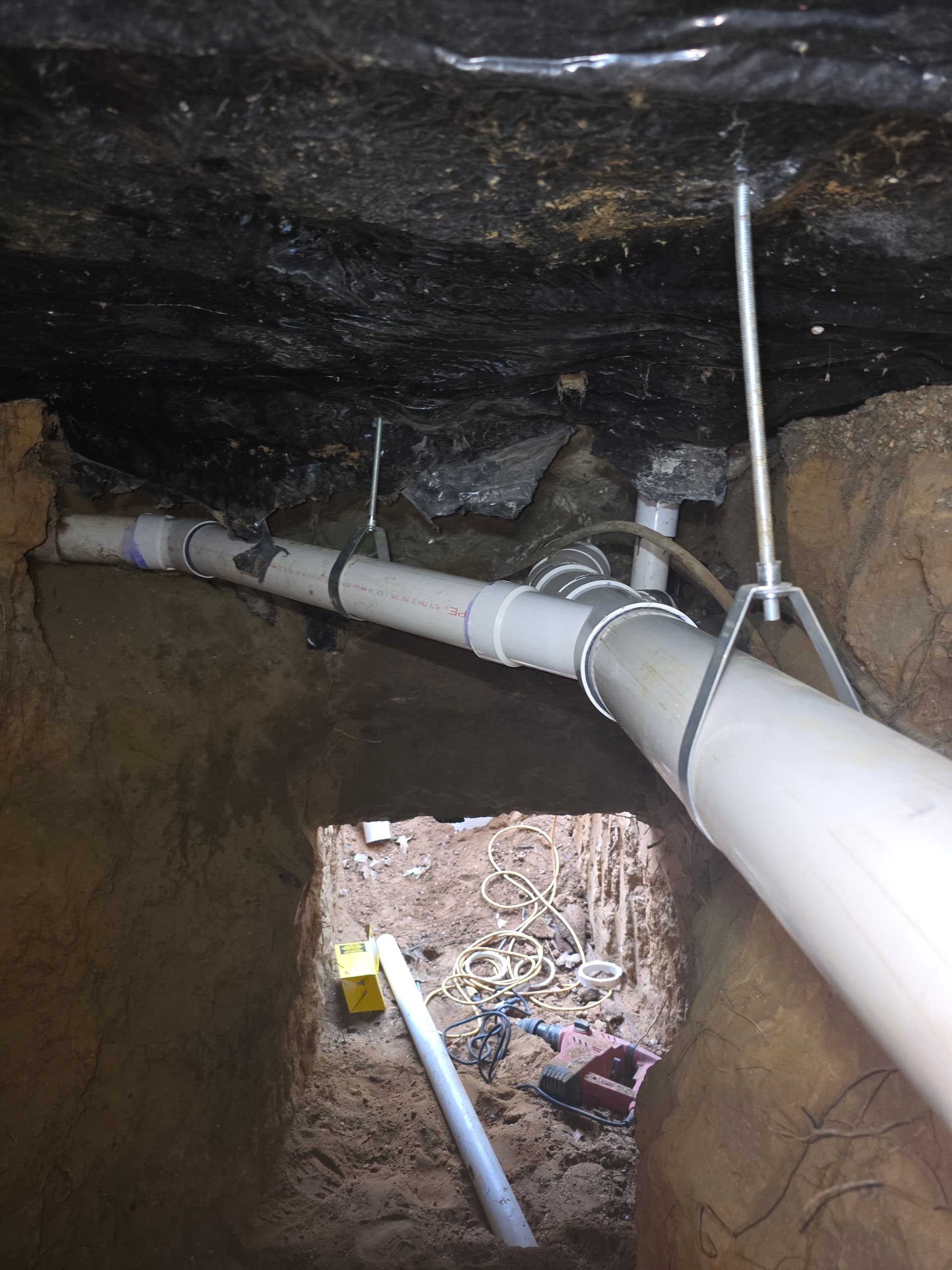 White PVC pipes installed in a crawlspace, secured by metal hangers. Dirt walls and ceiling surround the pipes.