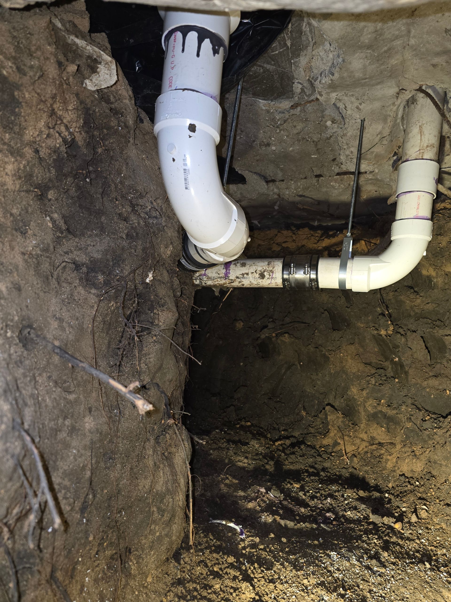 White PVC pipes connecting in a dark, dirt-filled space, likely a basement or crawlspace.