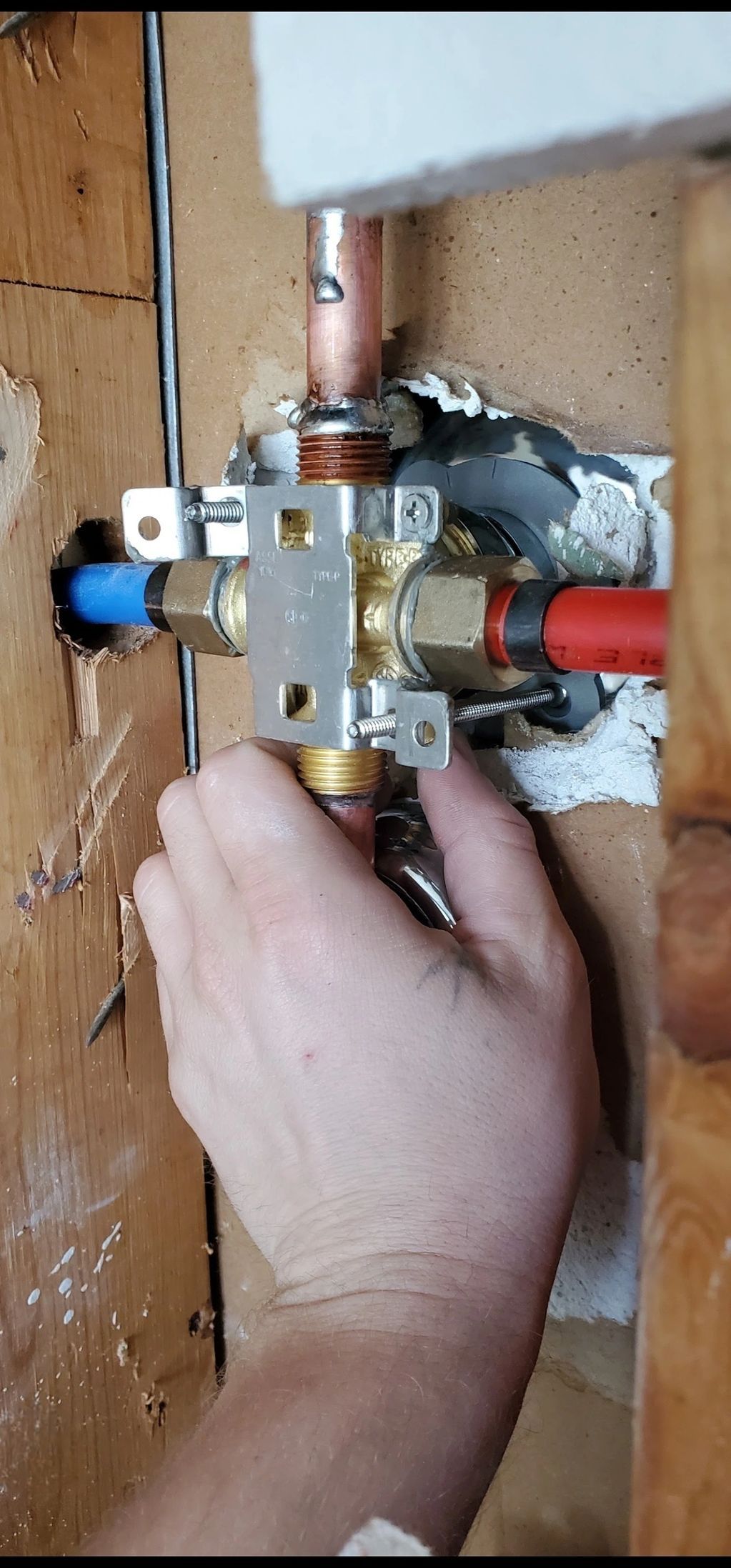 A hand turning a valve on a water pipe assembly inside a wooden wall.