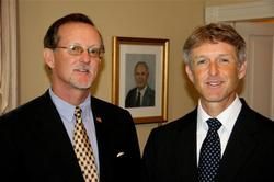 Two men in suits standing indoors, one in a striped tie and one in a dark tie, with a framed portrait behind them.