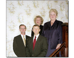 Four people posed on a staircase, with two men in front and two women behind, against floral wallpaper.