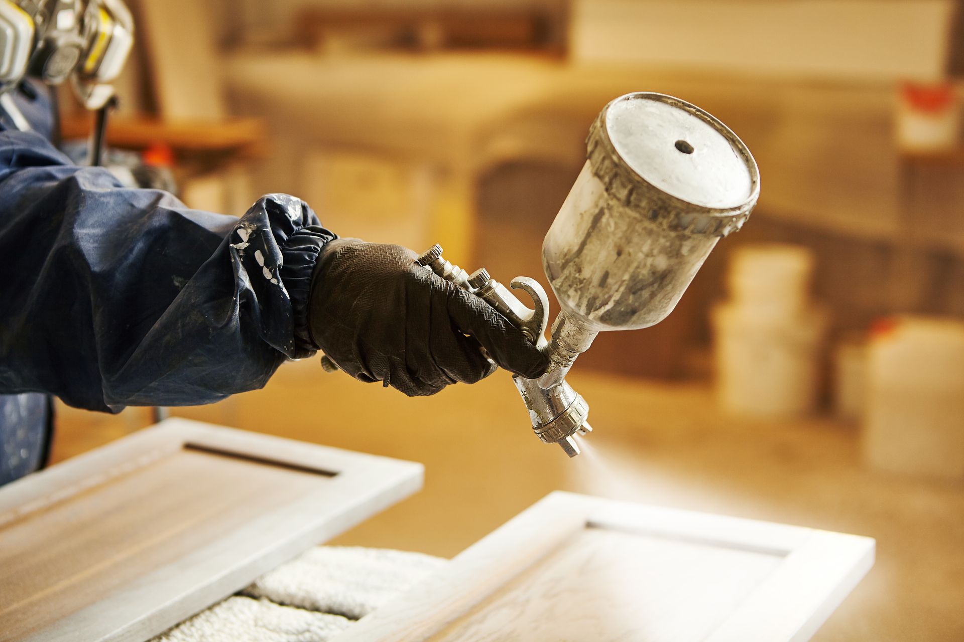 A person in a protective suit and respirator sprays a finish onto a wooden cabinet door with a paint gun.