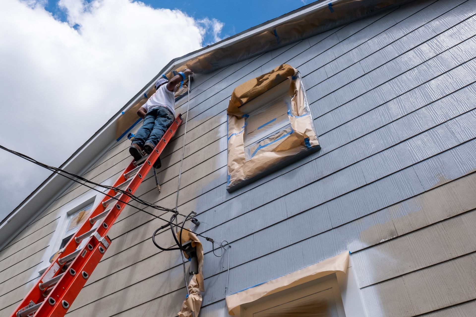 A person on an extension ladder paints the exterior siding of a two-story house under a cloudy blue sky.