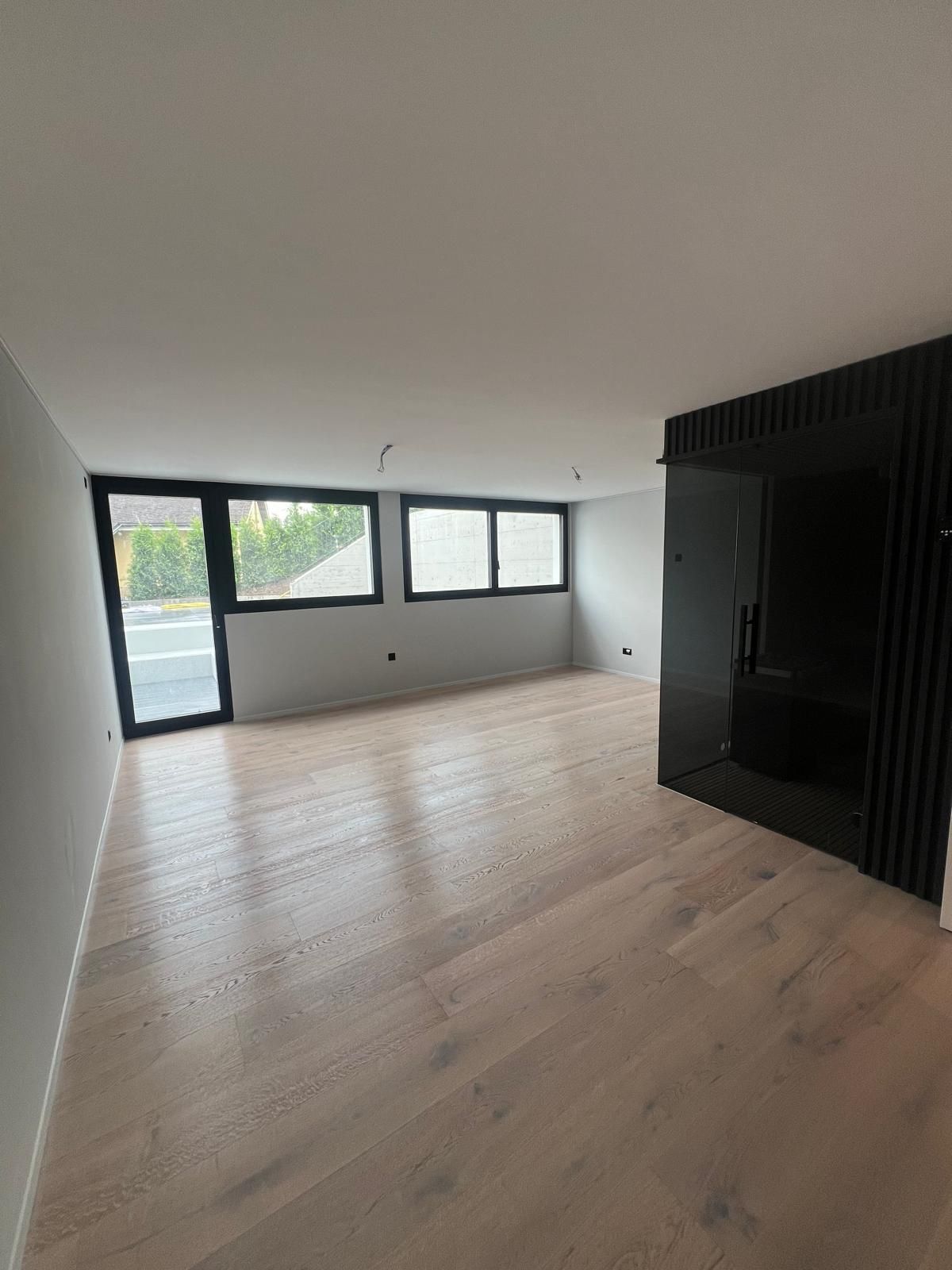 Leeres Zimmer mit hellen Holzböden, großen Fenstern und einer schwarzen Akzentwand.