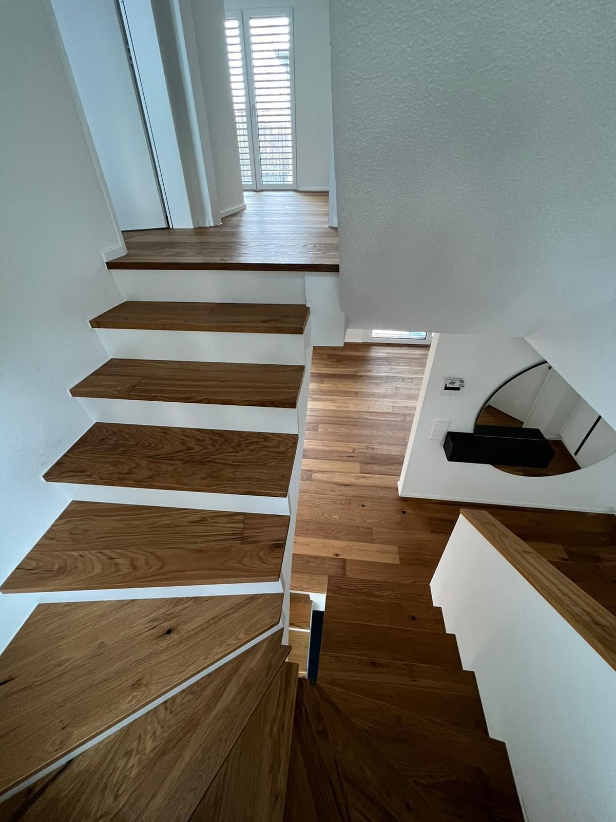 Eine Holztreppe mit weißen Wänden führt hinauf in einen hellen Flur und hinunter zu einem Treppenabsatz.