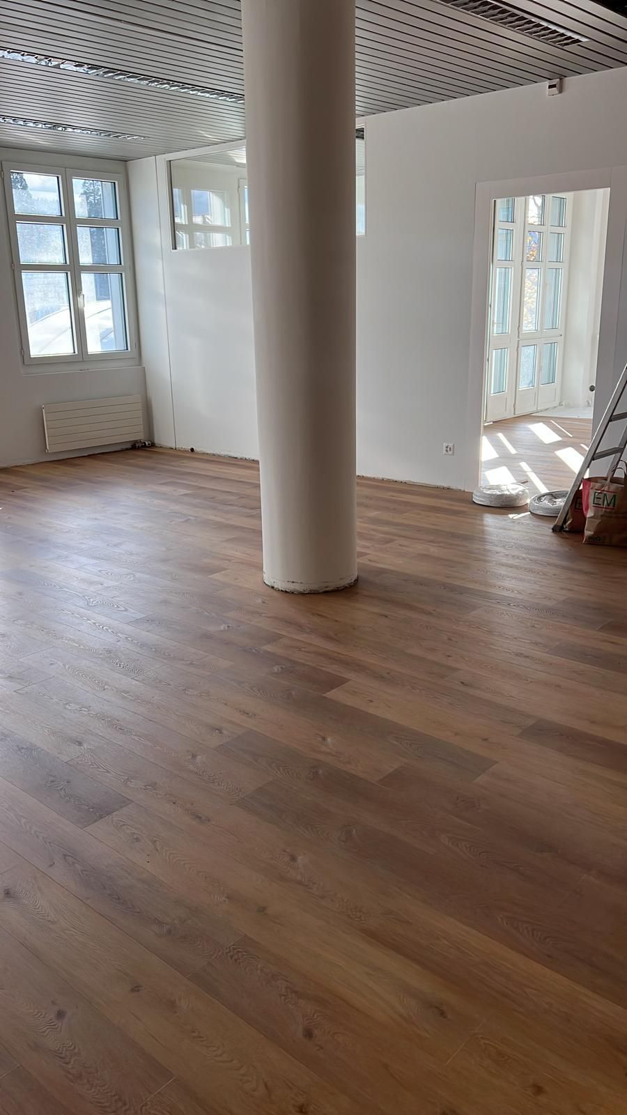 Ein leerer Raum mit Holzboden, weißen Wänden und einer großen zentralen Säule. Natürliches Licht strömt herein.