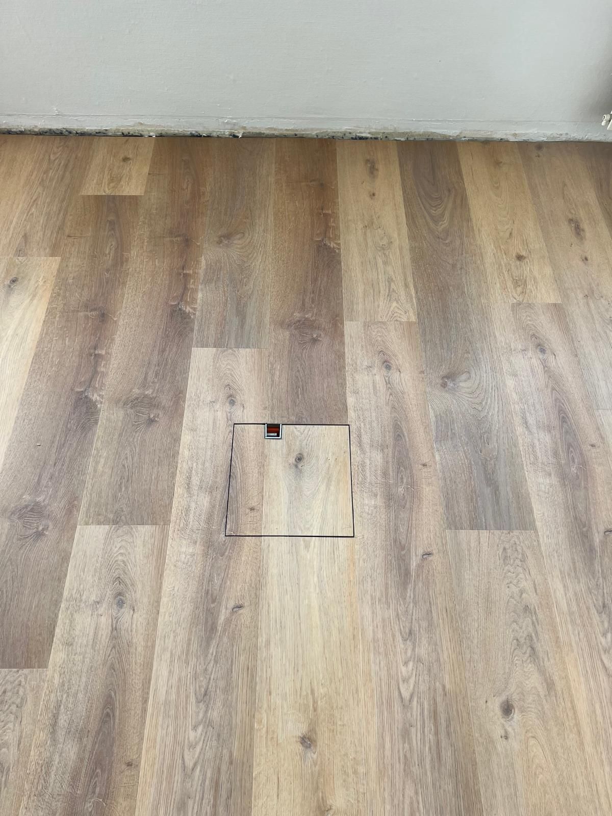 Beschädigter Holzboden mit einem quadratischen Loch. Der Boden ist hellbraun und grenzt an eine Wand.