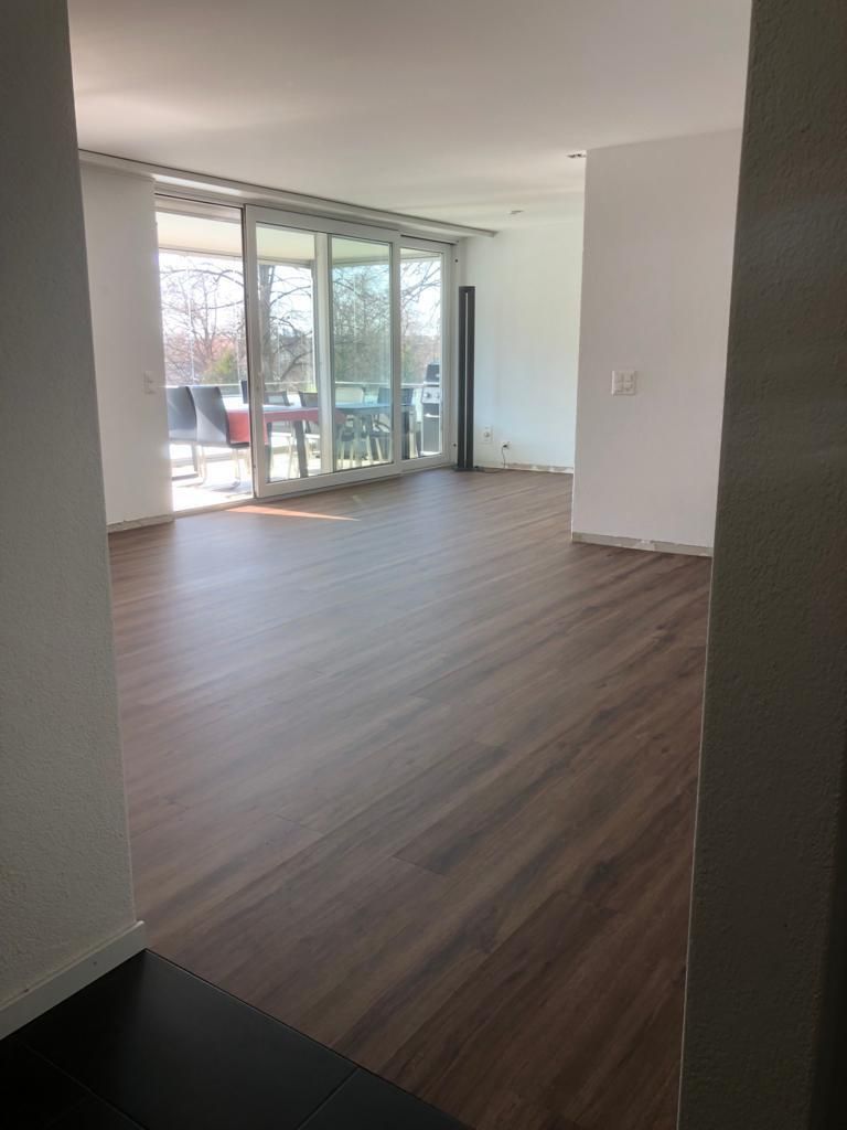 Leeres Wohnzimmer mit Holzboden, weißen Wänden und Glastüren, die auf einen Balkon führen.