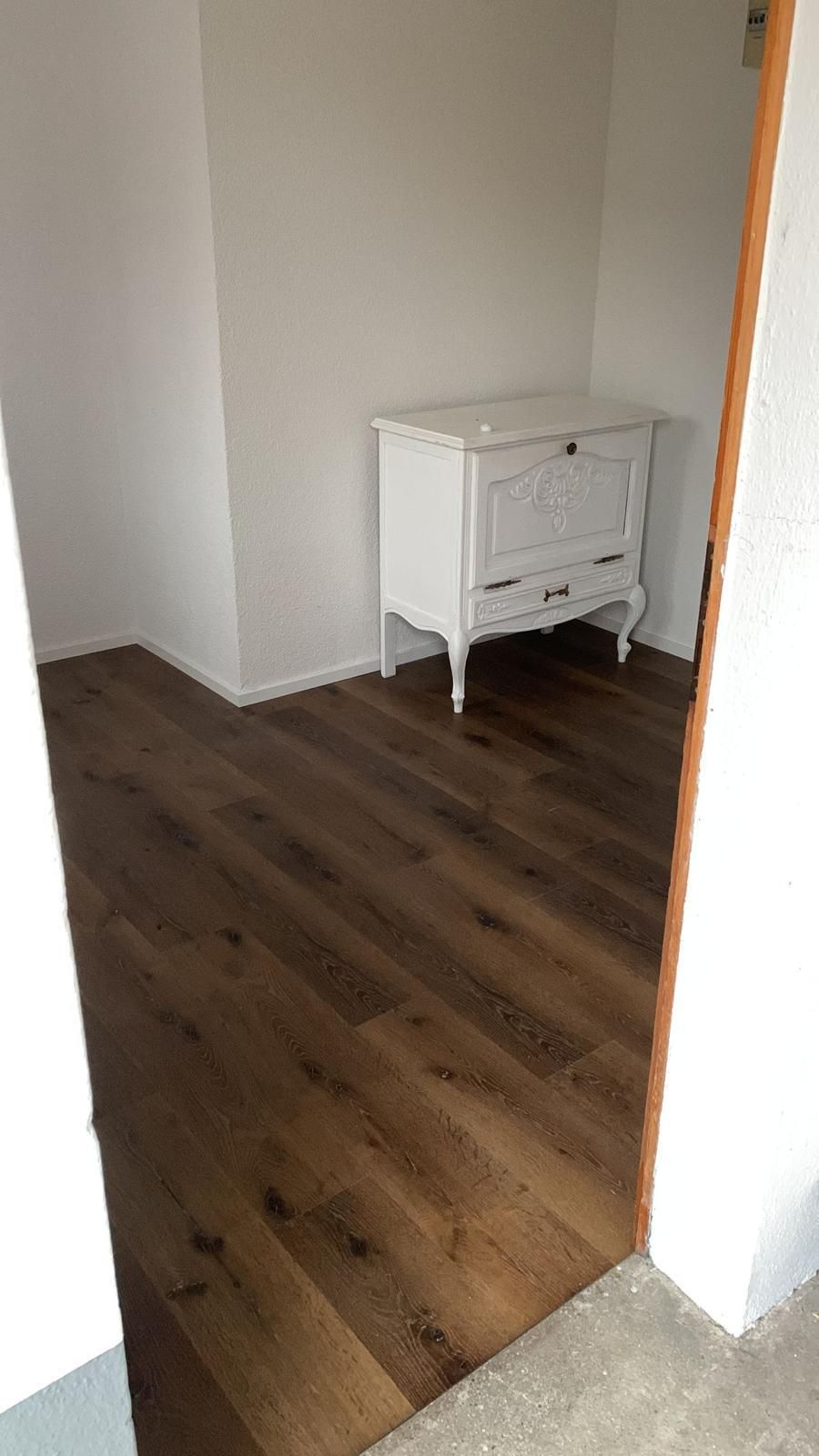 Zimmer mit dunklem Holzboden, weißer Kommode vor weißen Wänden, Tür auf der rechten Seite.