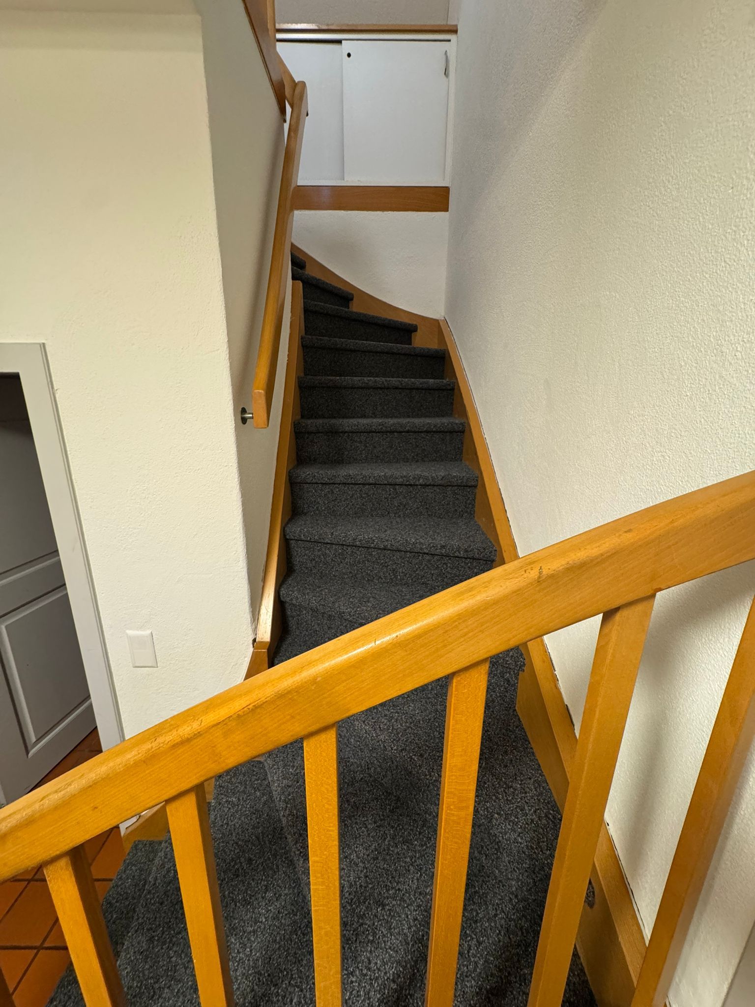 Treppe mit grauem Teppichboden und Holzhandlauf, die zu einem Treppenabsatz mit einem Einbauschrank und weißen Wänden führt.