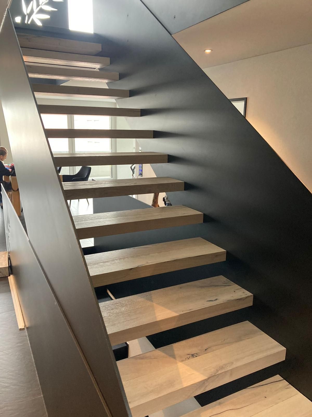 Moderne Treppe mit freischwebenden Holzstufen, schwarzer Metallstütze und einer Person im Hintergrund.