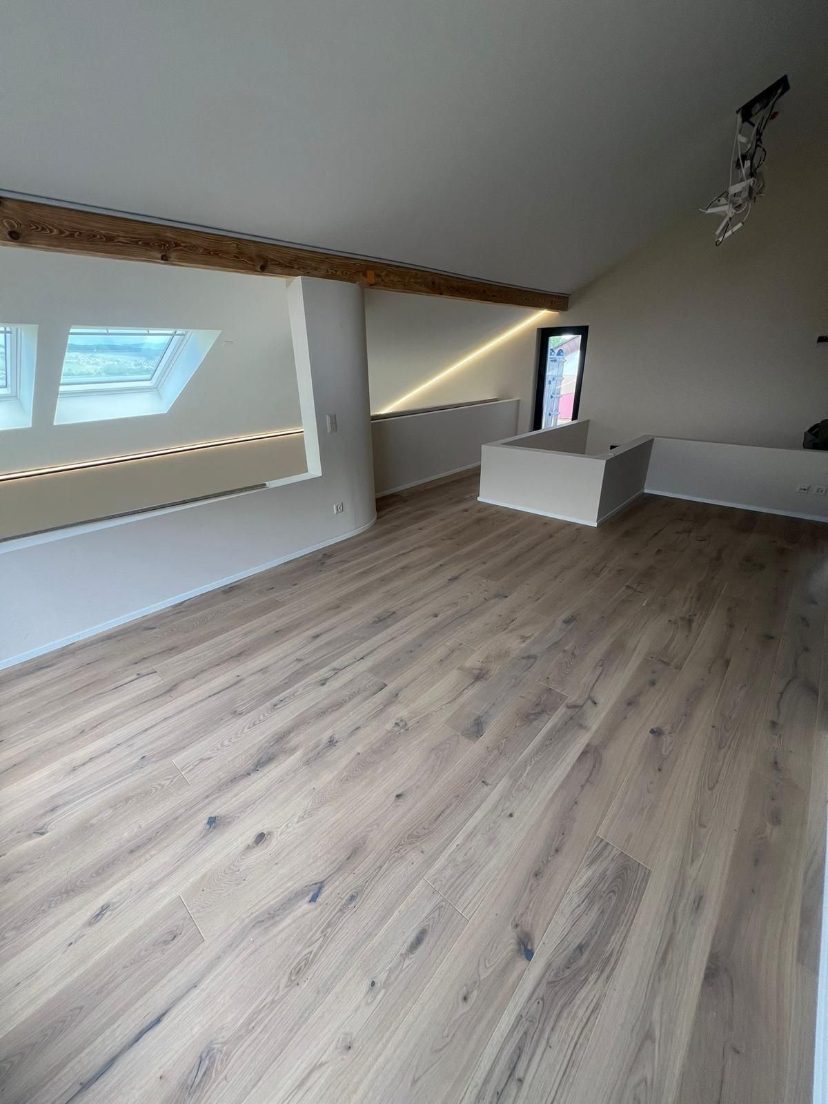 Zimmer mit Holzbodenoptik, schräger Decke mit Oberlicht und Holzbalken sowie eingebautem weißen Ablagebrett.
