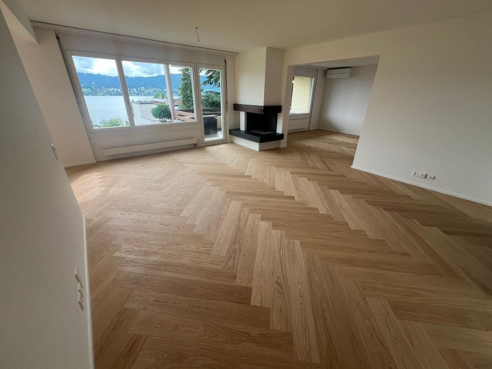 Heller Innenraum mit Fischgrätparkett, Kamin und großen Fenstern mit Blick aufs Wasser.