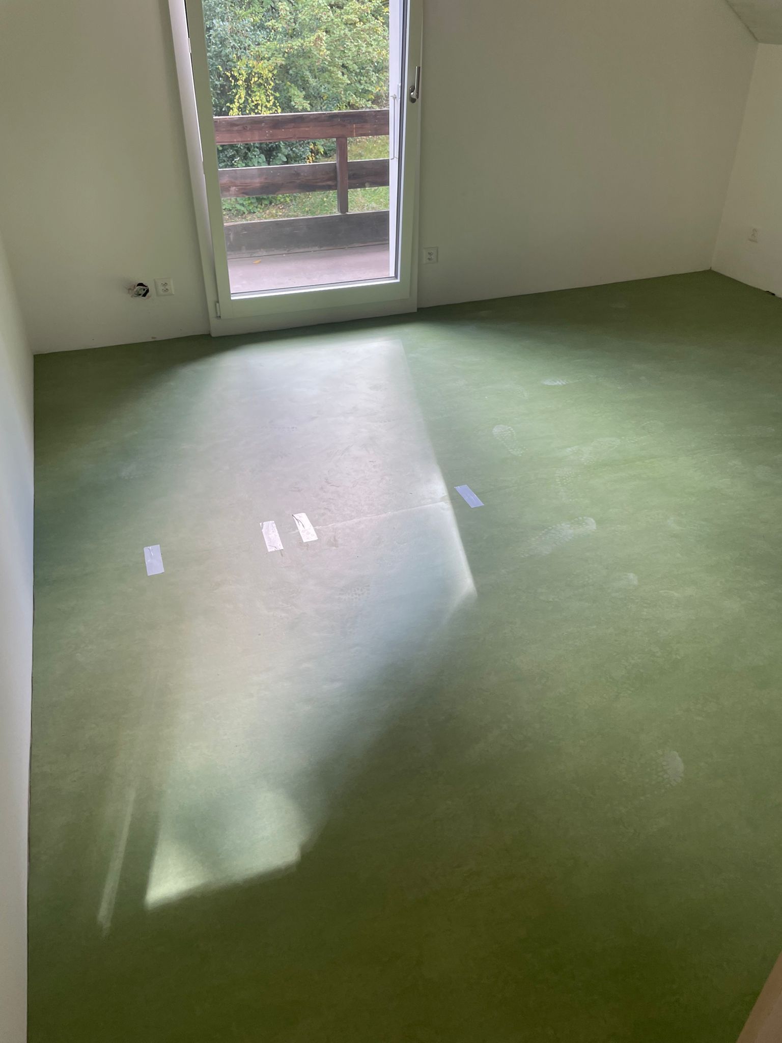 Leerer Raum mit grünem Boden und einer Tür, die auf einen Balkon führt. Sonnenlicht strömt über den Boden.