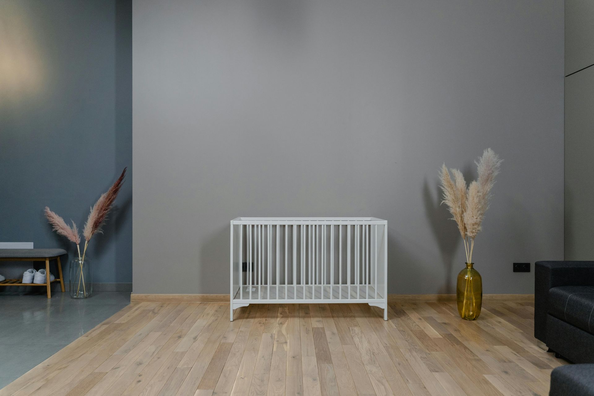 Weißes Kinderbett in einem Zimmer mit grauen Wänden und Holzboden, flankiert von Vasen mit Pampasgras.
