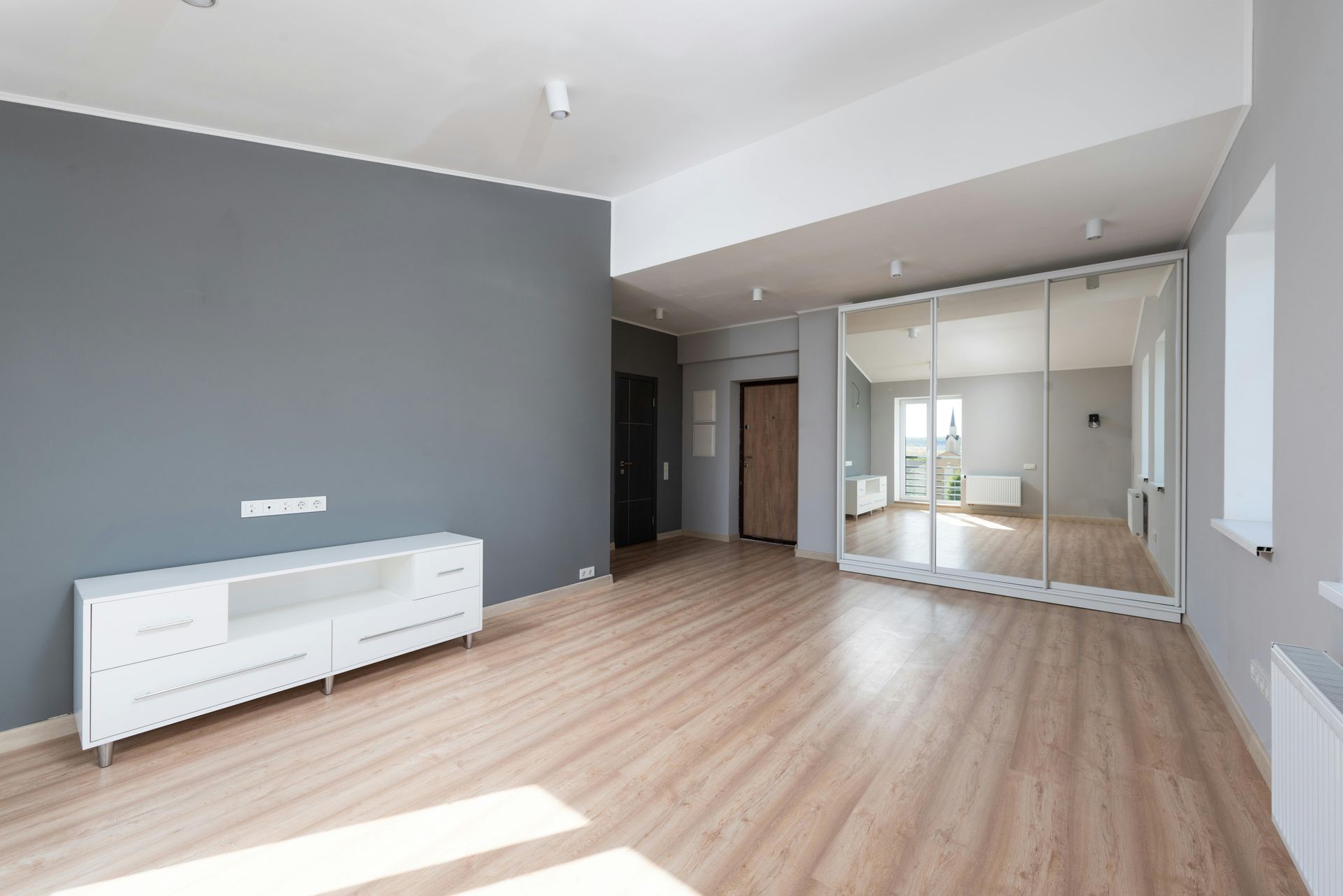 Leeres Zimmer mit grauer Akzentwand, weißem Aufbewahrungsschrank, hellem Holzboden und Spiegelschrank.