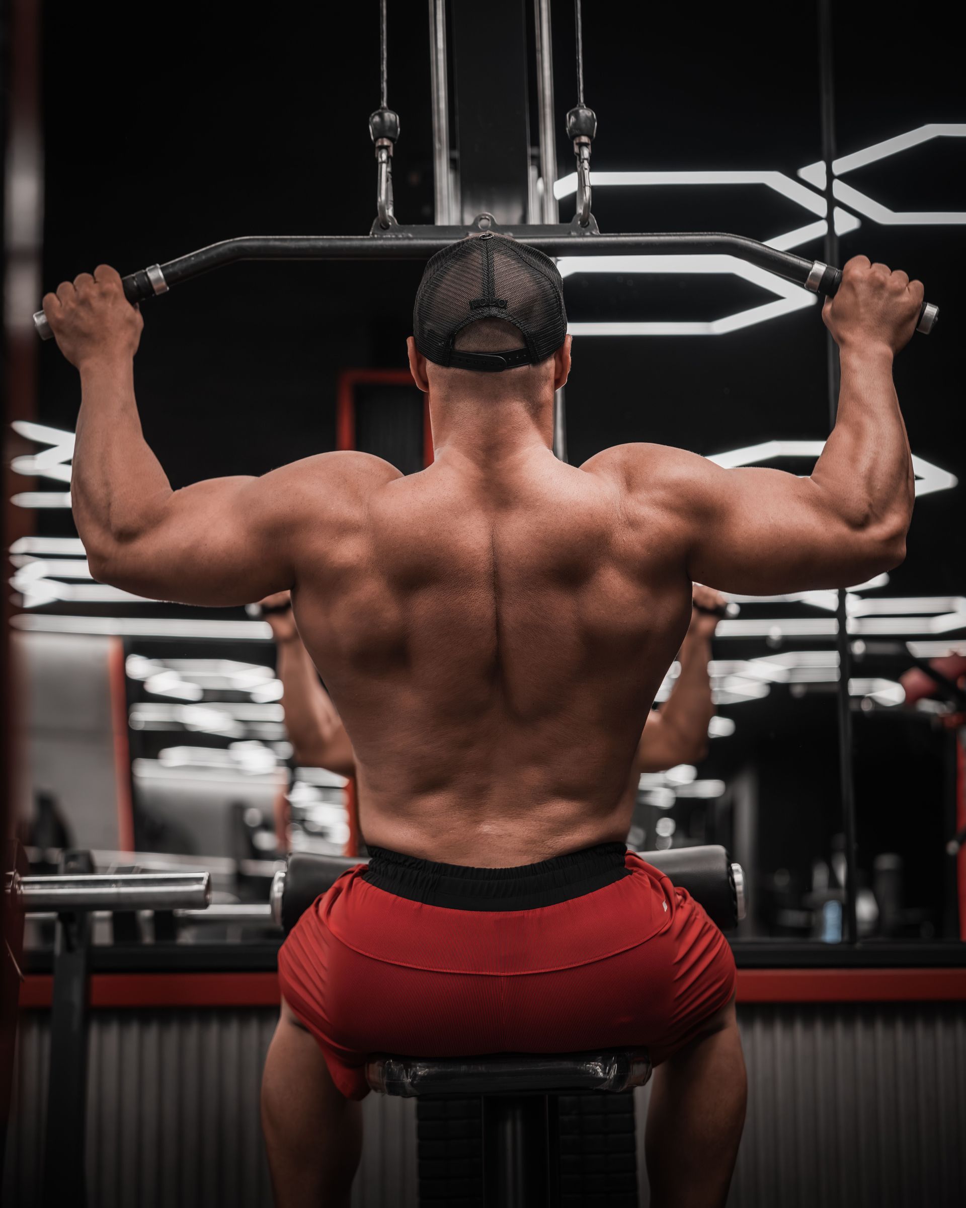 Muscular man doing a lat pulldown in a gym, seen from behind, wearing red shorts.