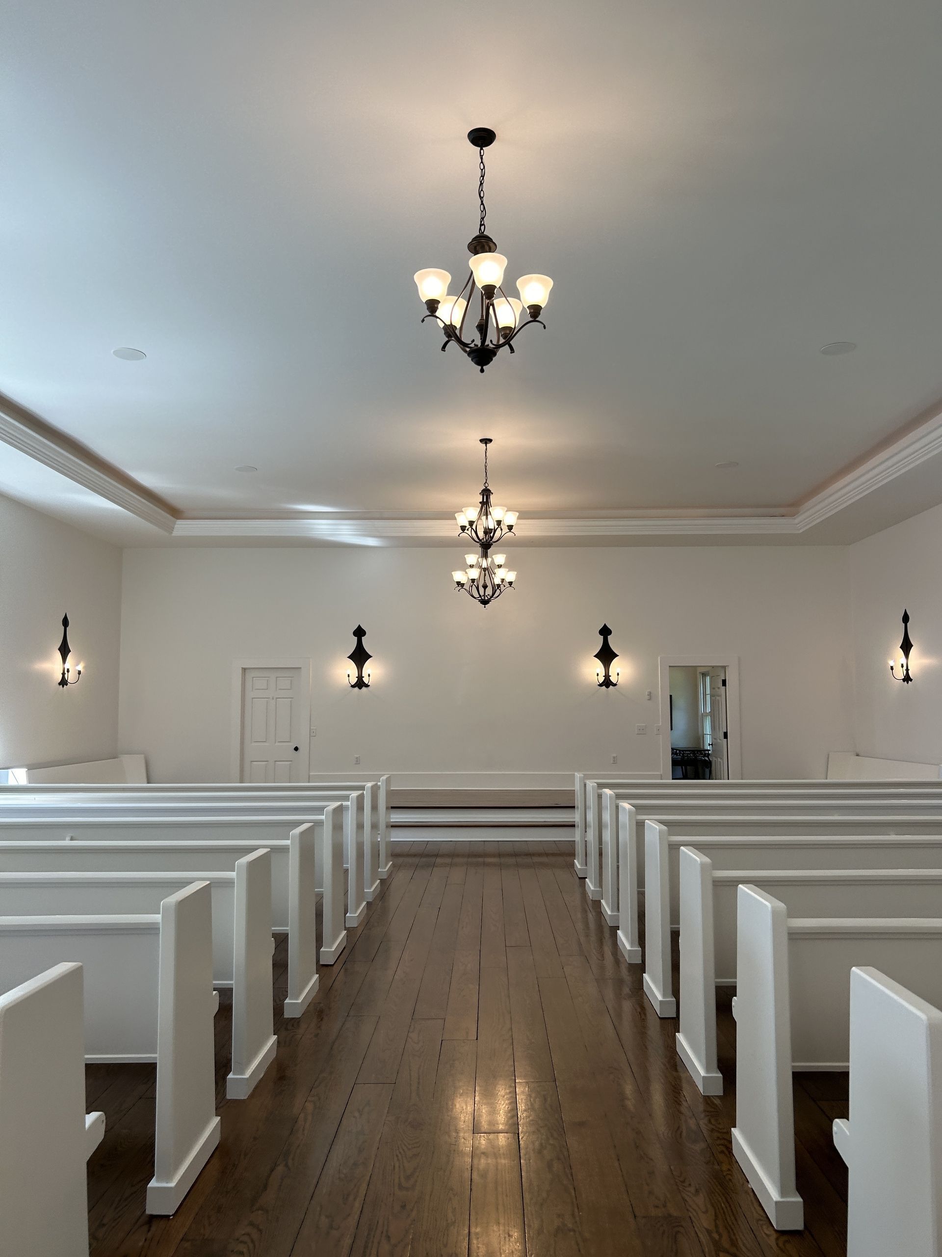 An aisle between white pews in a chapel with wooden floors, white walls, and ornate chandeliers overhead.
