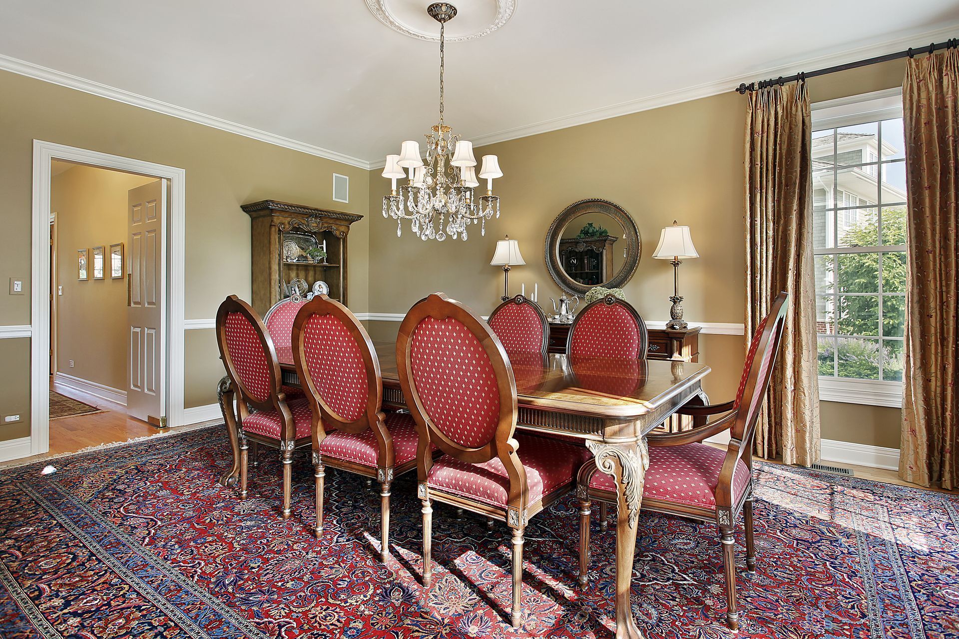 Formal dining room with a mahogany table, six red patterned chairs, crystal chandelier, and a large oriental rug.