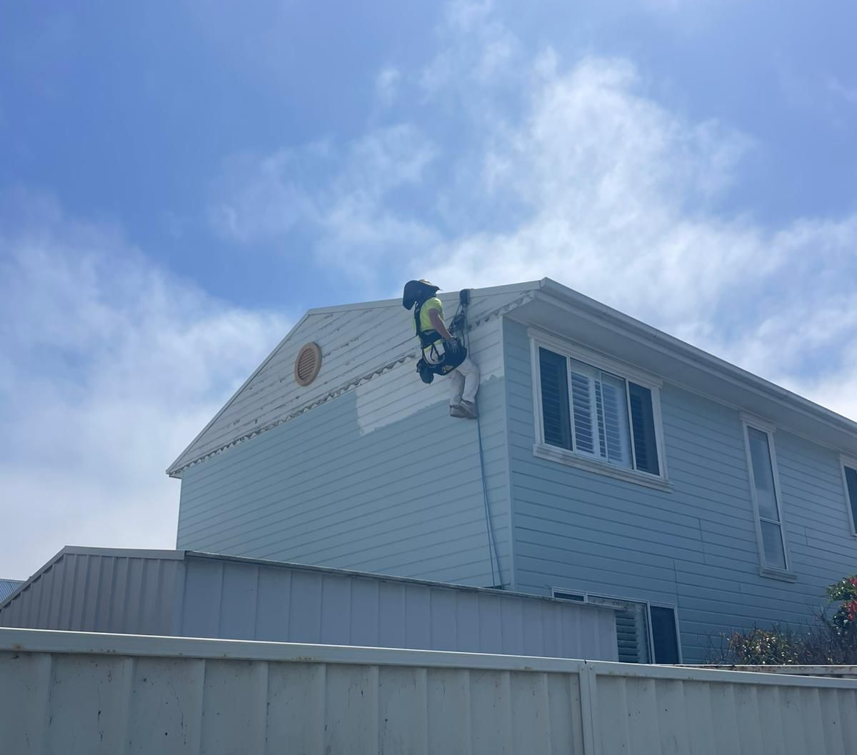 Person in safety harness painting the side of a blue house, blue sky in background.