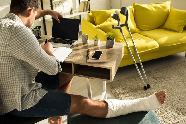Man in a cast using a laptop by a coffee table, with crutches and a yellow sofa in the background