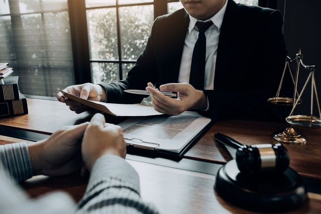 Lawyer in a dark suit reviewing documents across a desk with a client, gavel and scales nearby