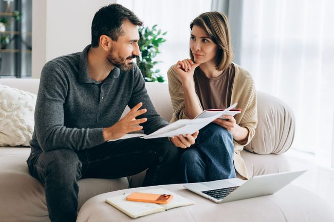 Two people discussing papers on a couch beside a laptop in a bright living room