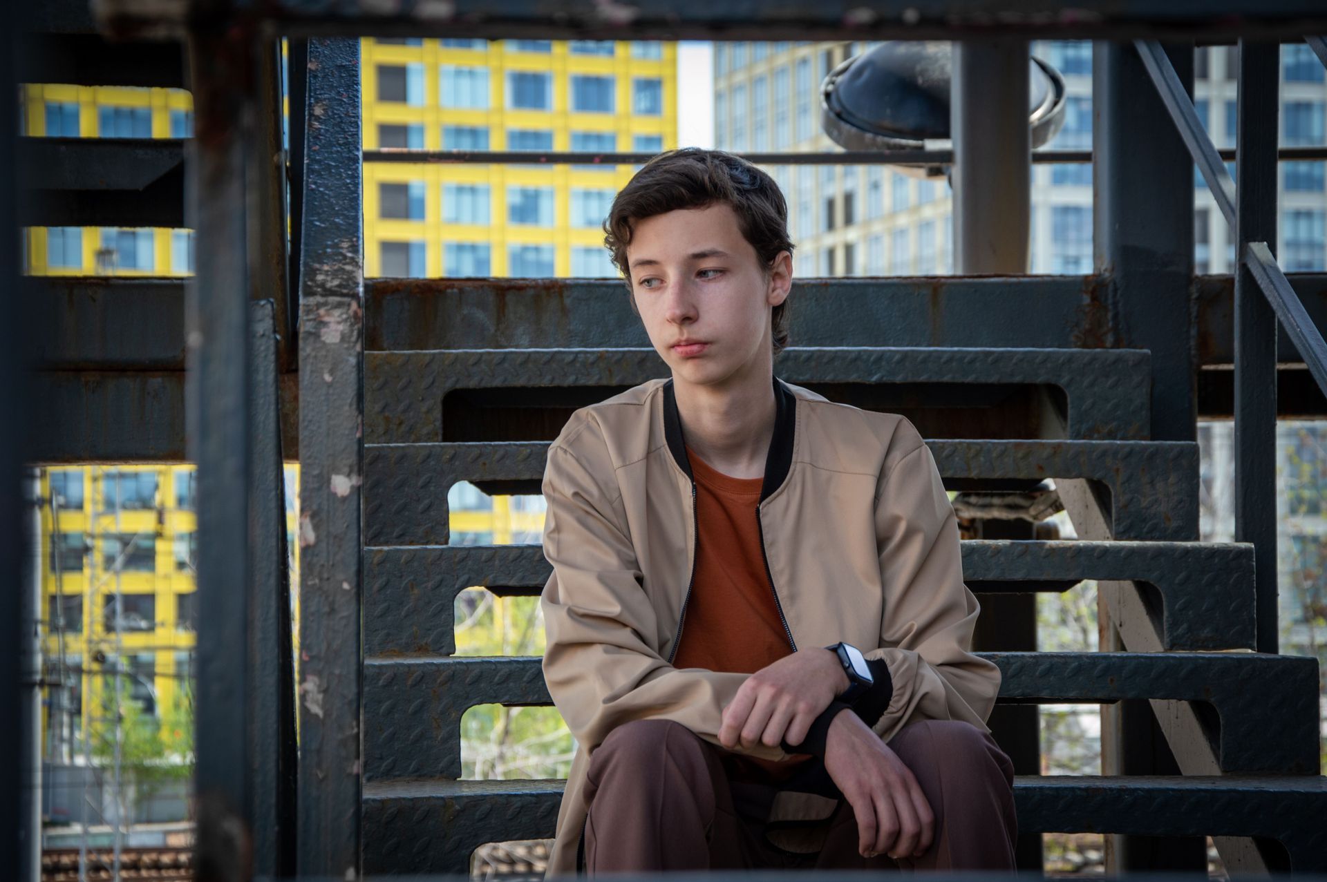 Young man sitting on outdoor metal stairs in a city setting, wearing a tan jacket and rust-colored shirt.