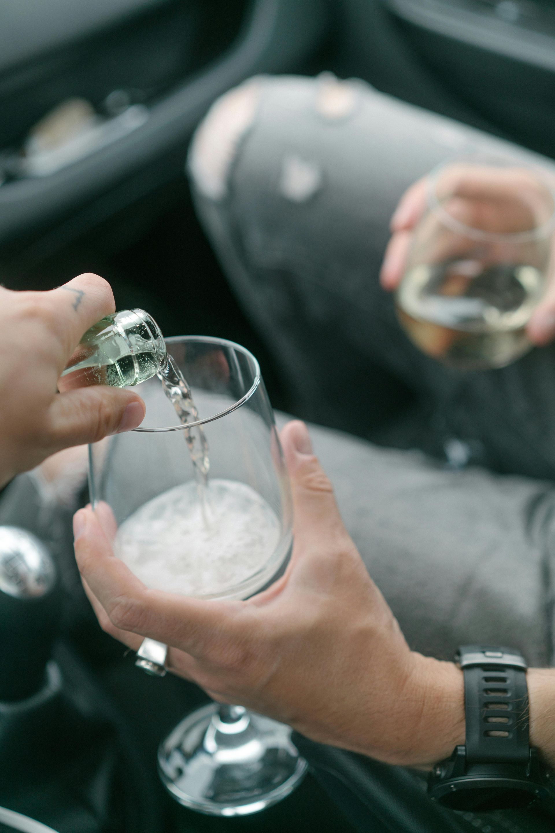 Hands pouring a clear drink into a champagne flute while another glass is held nearby in a car interior