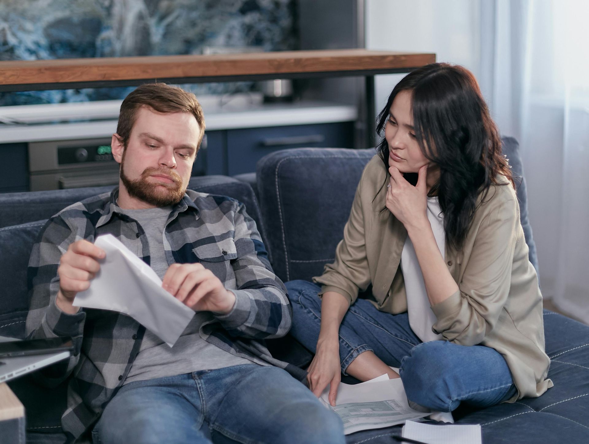 Two people sit on a couch, looking at papers and talking in a living room.