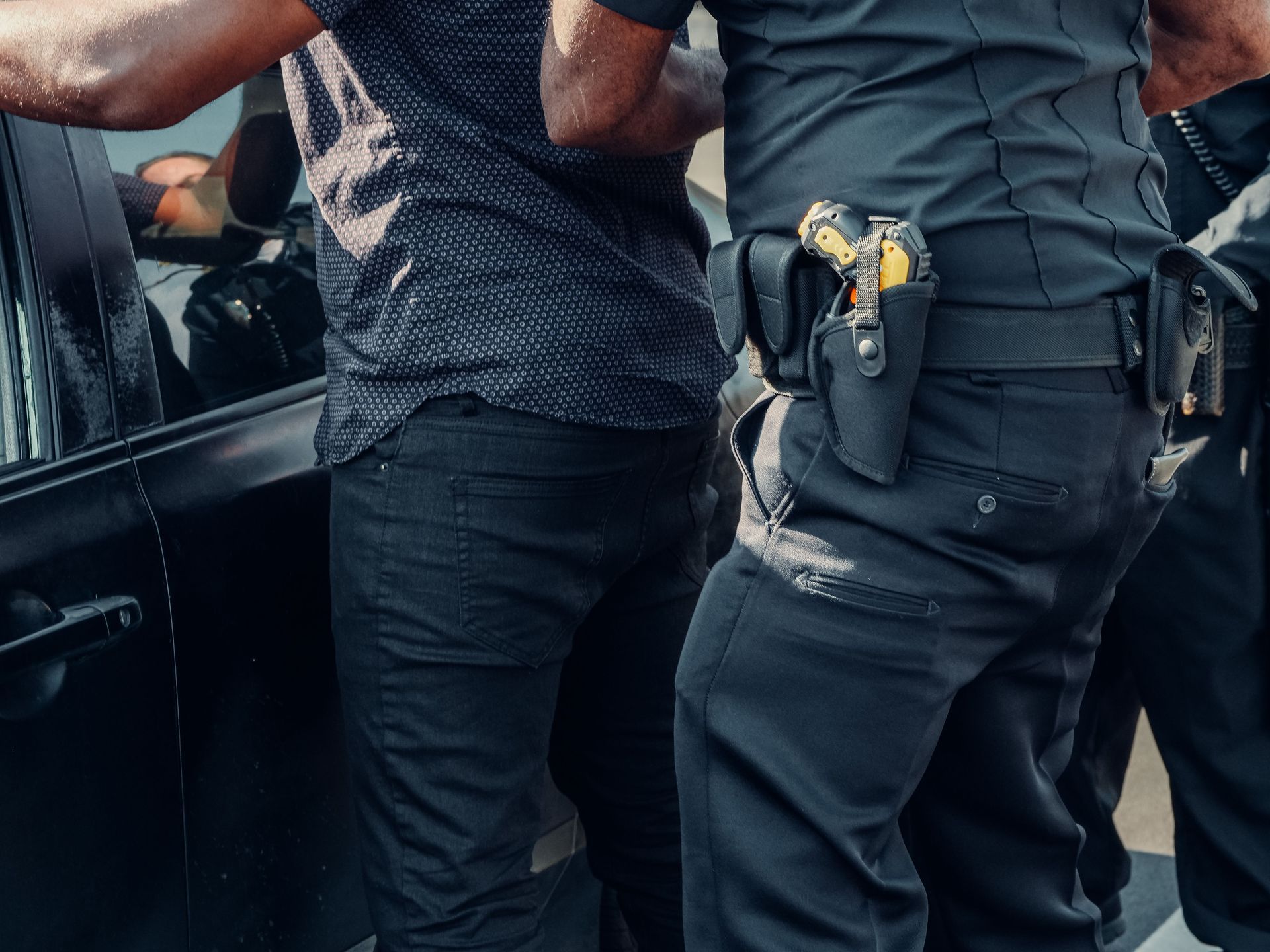 Two officers detain a person beside a car, with one officer’s hand on the person’s arm.