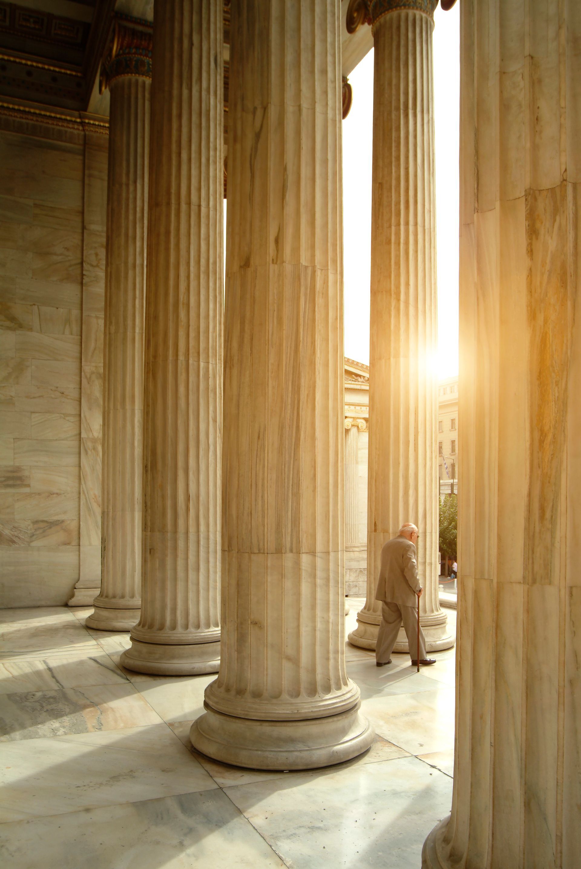 Sunlit classical columns framing a person walking through a stone colonnade