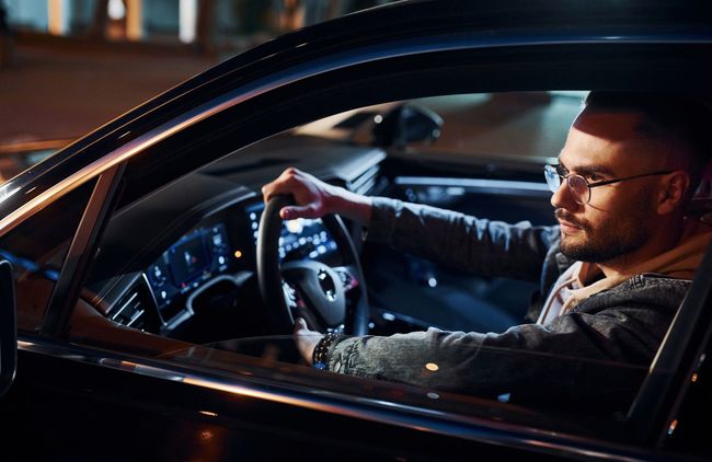 Man driving a car at night, lit by dashboard lights and city reflections through the window