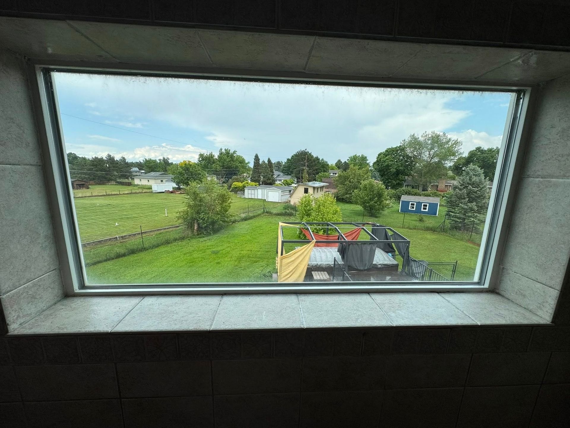 View through a window, looking out onto a green yard with trees and houses, under a cloudy sky.